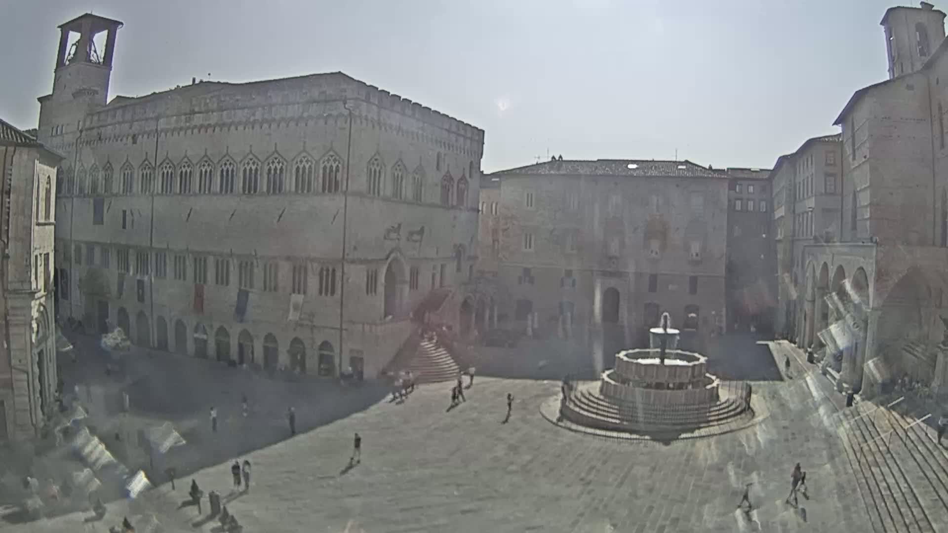 A sunny day reveals a large town square with a fountain in the center, surrounded by historic stone buildings.