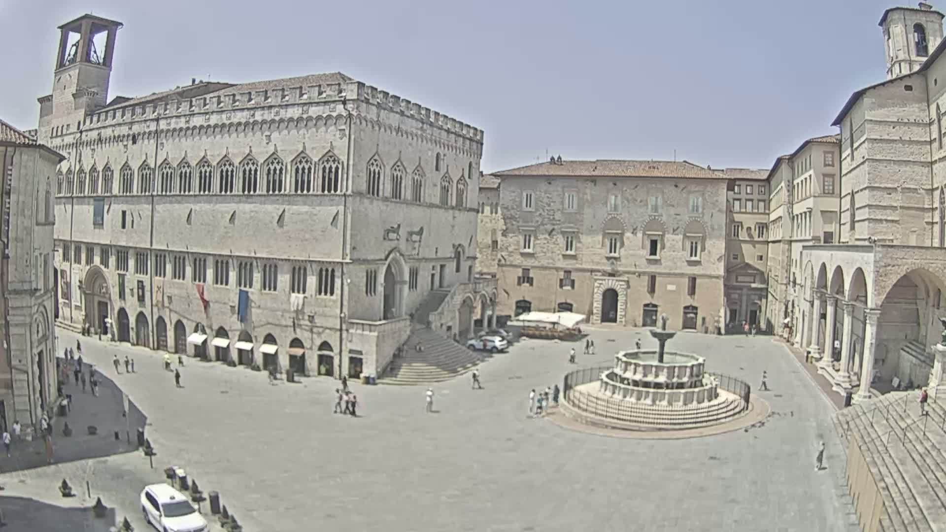 A large, sunny town square with a fountain in the center is surrounded by historical stone buildings.