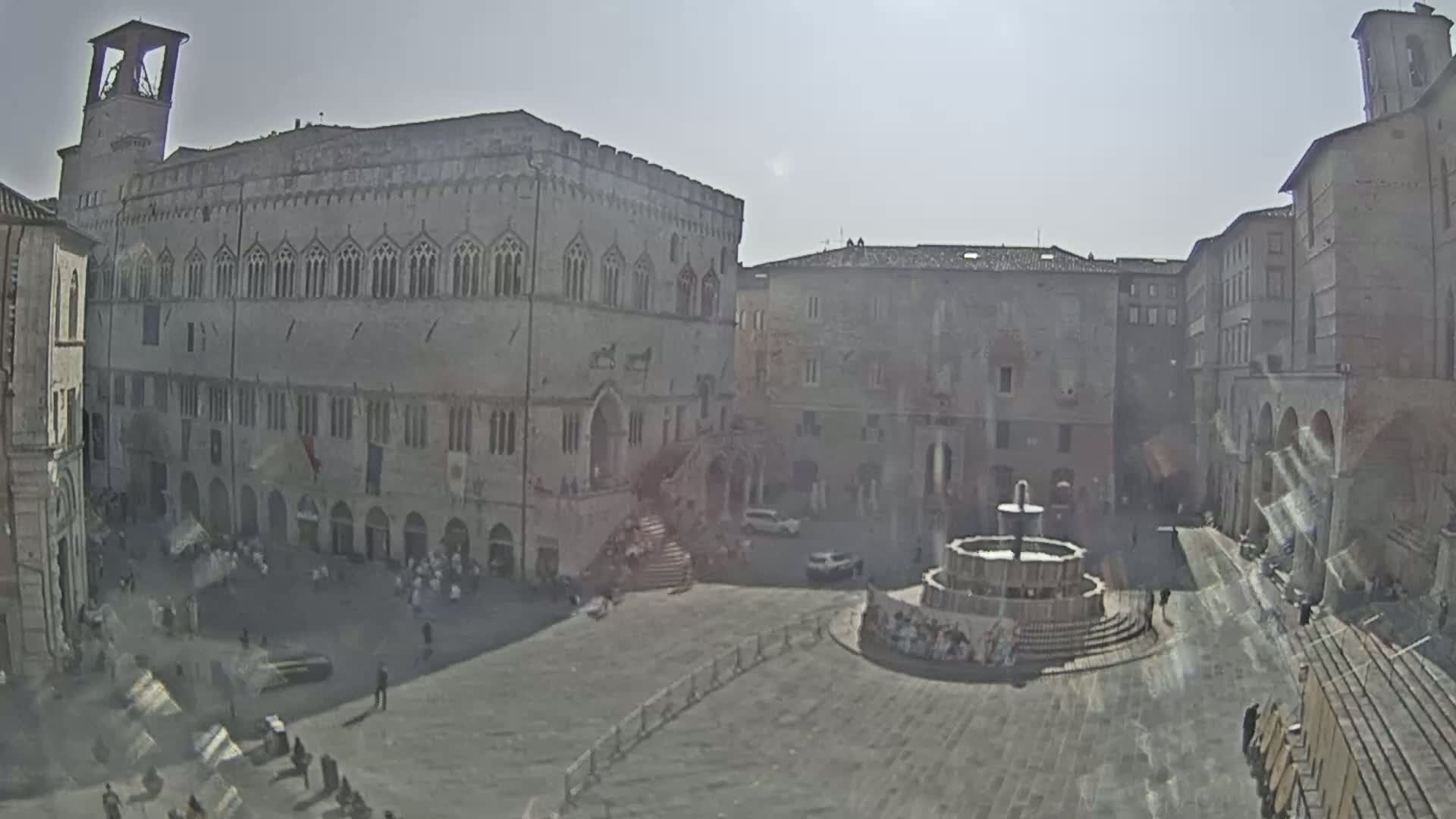A large, sunlit town square with a fountain at its center is surrounded by old stone buildings, one of which is a large palace with a bell tower.