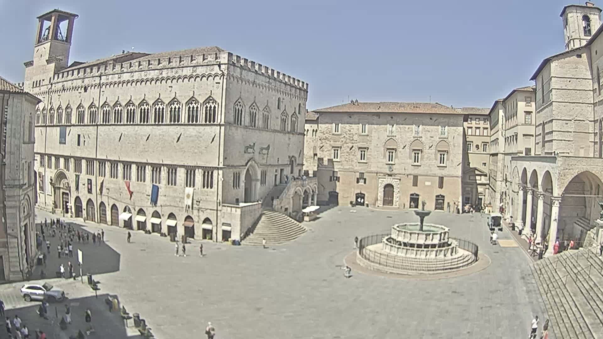 A large, sunny town square is surrounded by several stone buildings, one of which has a large staircase leading to its entrance, and a fountain sits in the center.