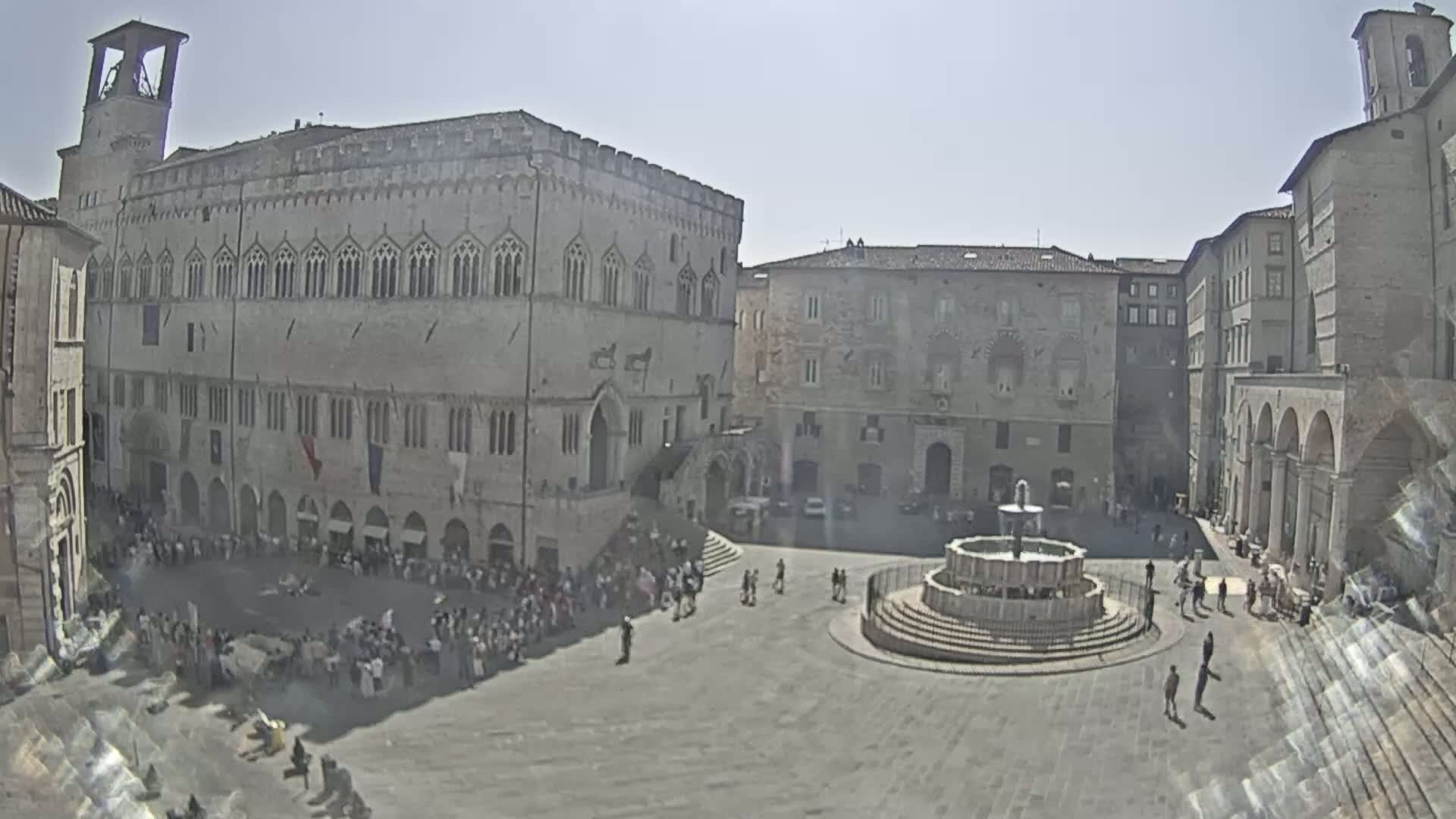 A large town square, under sunny skies, is filled with people and features a central fountain surrounded by medieval-style buildings.