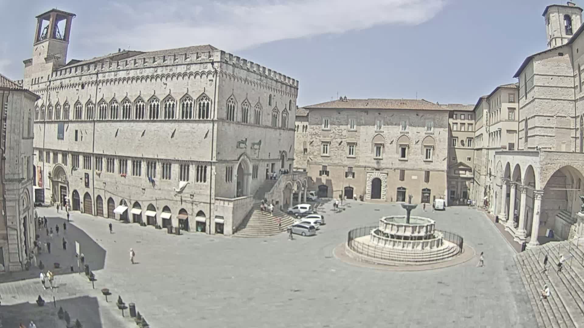 A large, light-colored stone plaza with a fountain in the center is surrounded by historic buildings under a partly cloudy sky.