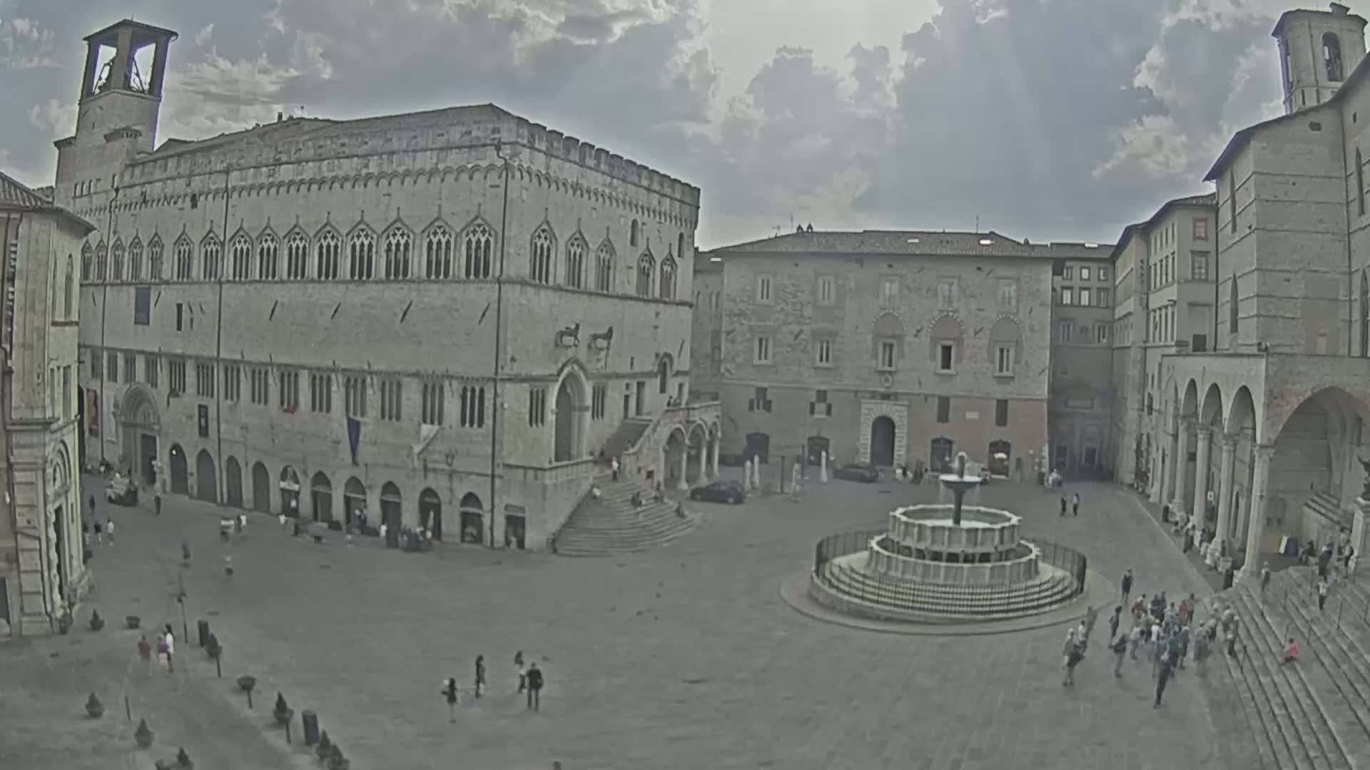 A large town square with a fountain at its center, surrounded by several stone buildings under a cloudy sky.