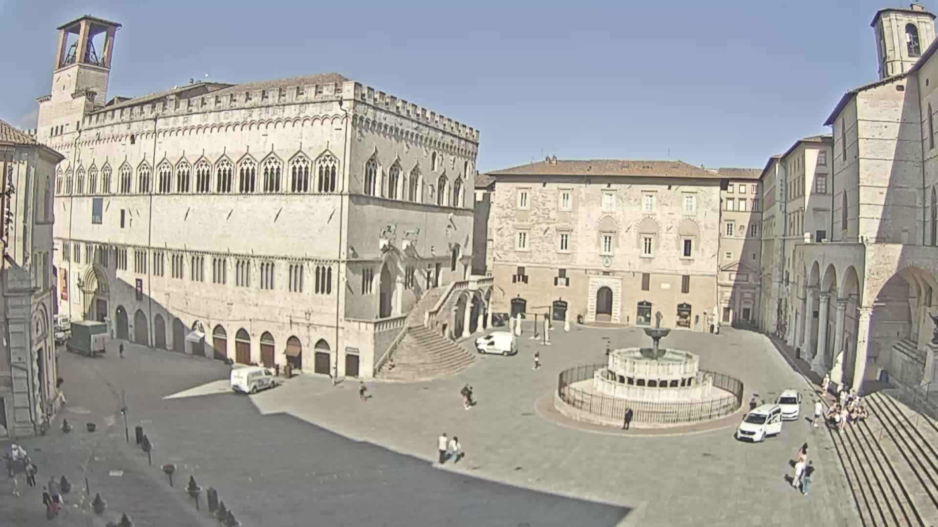 A large, mostly empty town square on a sunny day features a central fountain, surrounded by historic stone buildings.
