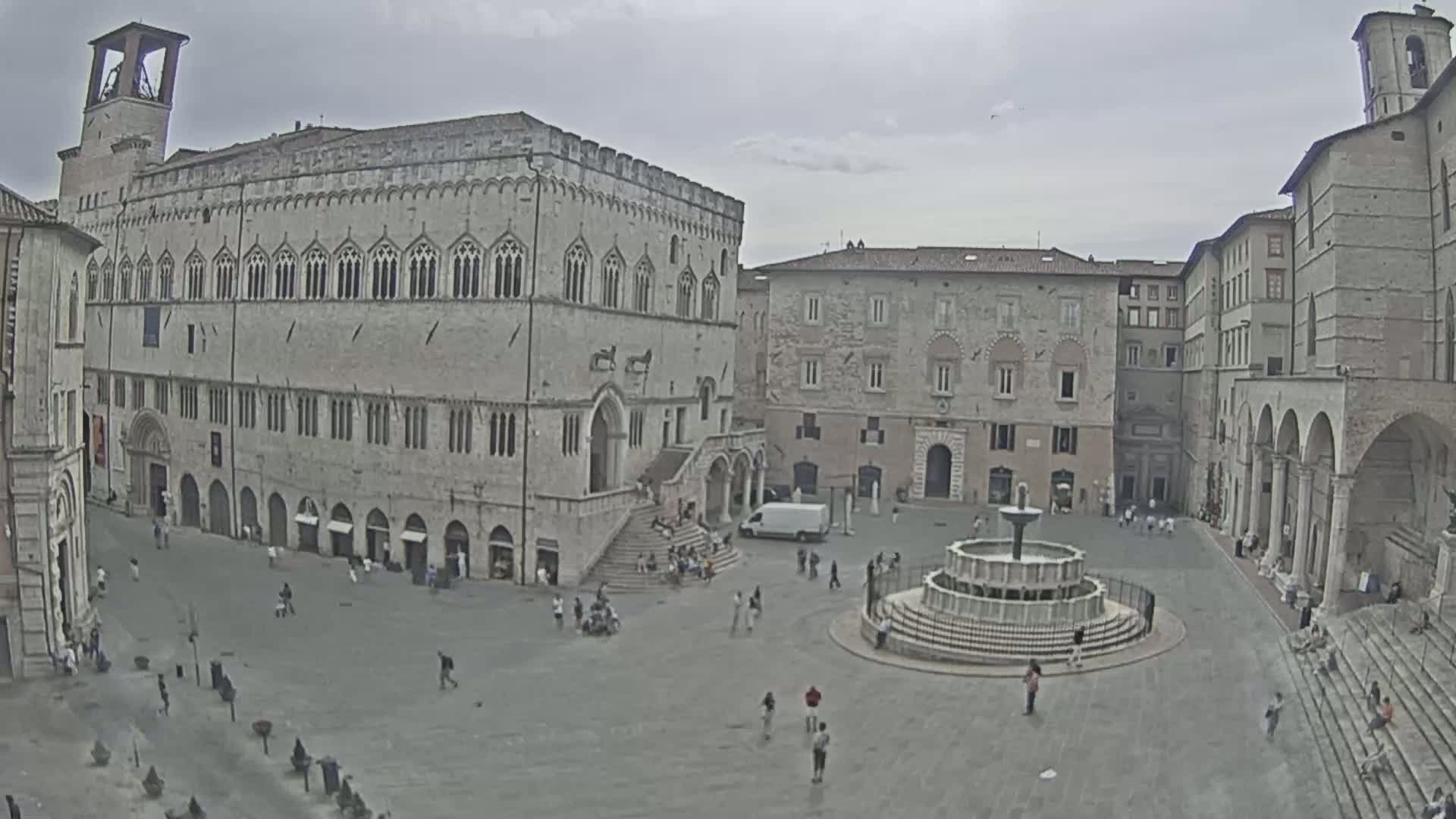 A large town square with several stone buildings surrounding it, a fountain in the center, and a few people scattered throughout under an overcast sky.