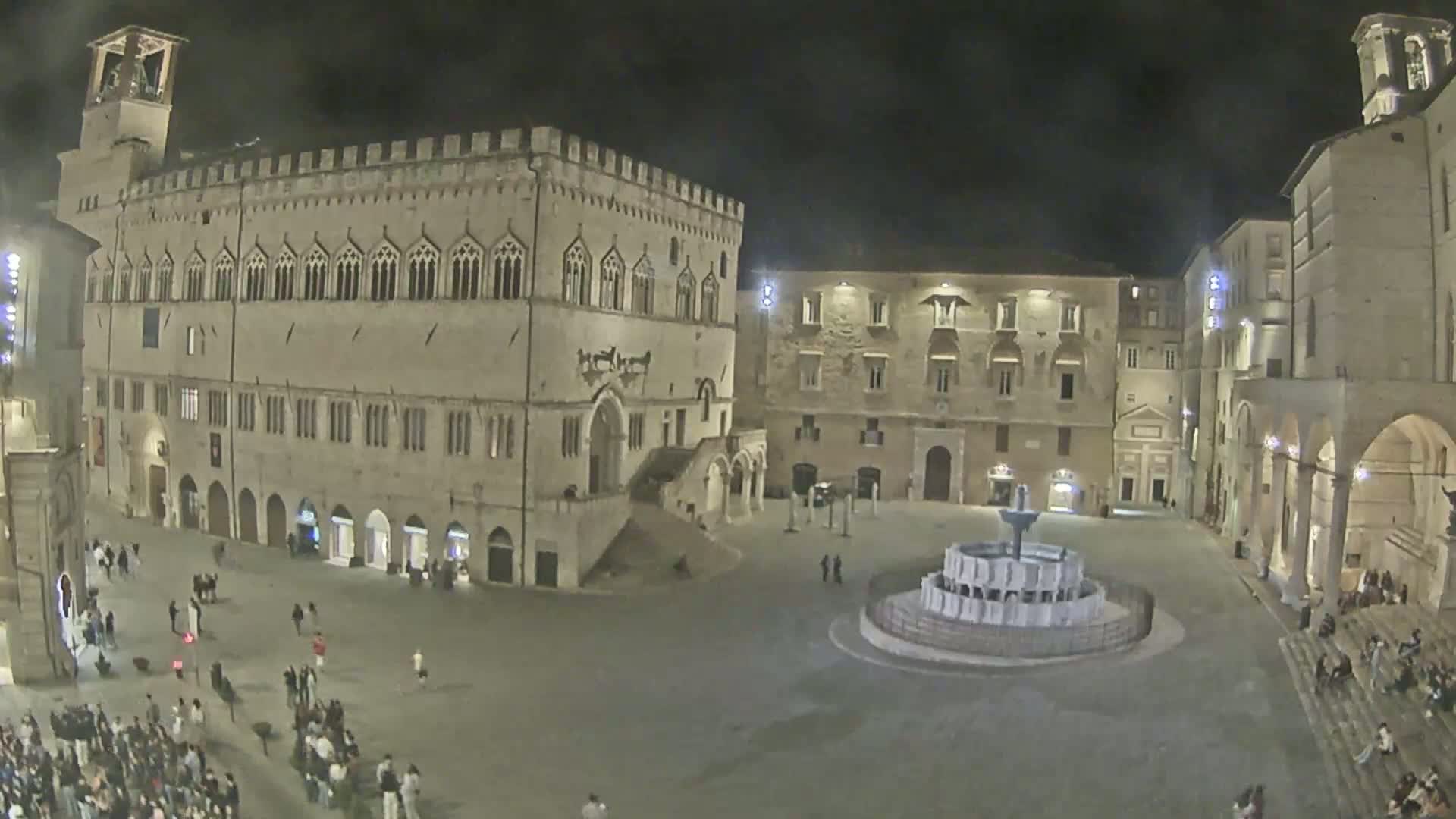 A large, illuminated town square at night, featuring a central fountain, and surrounded by historic stone buildings.