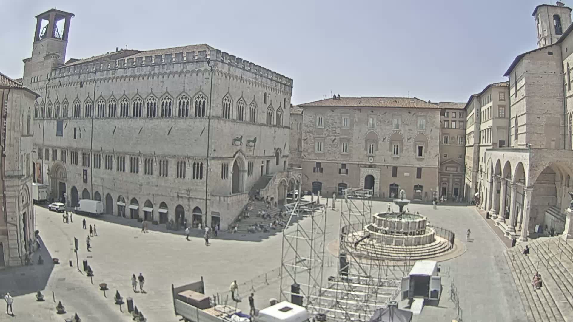 A large town square, under sunny skies, is surrounded by historic stone buildings, one prominently featuring a large staircase and a central fountain with scaffolding surrounding it.