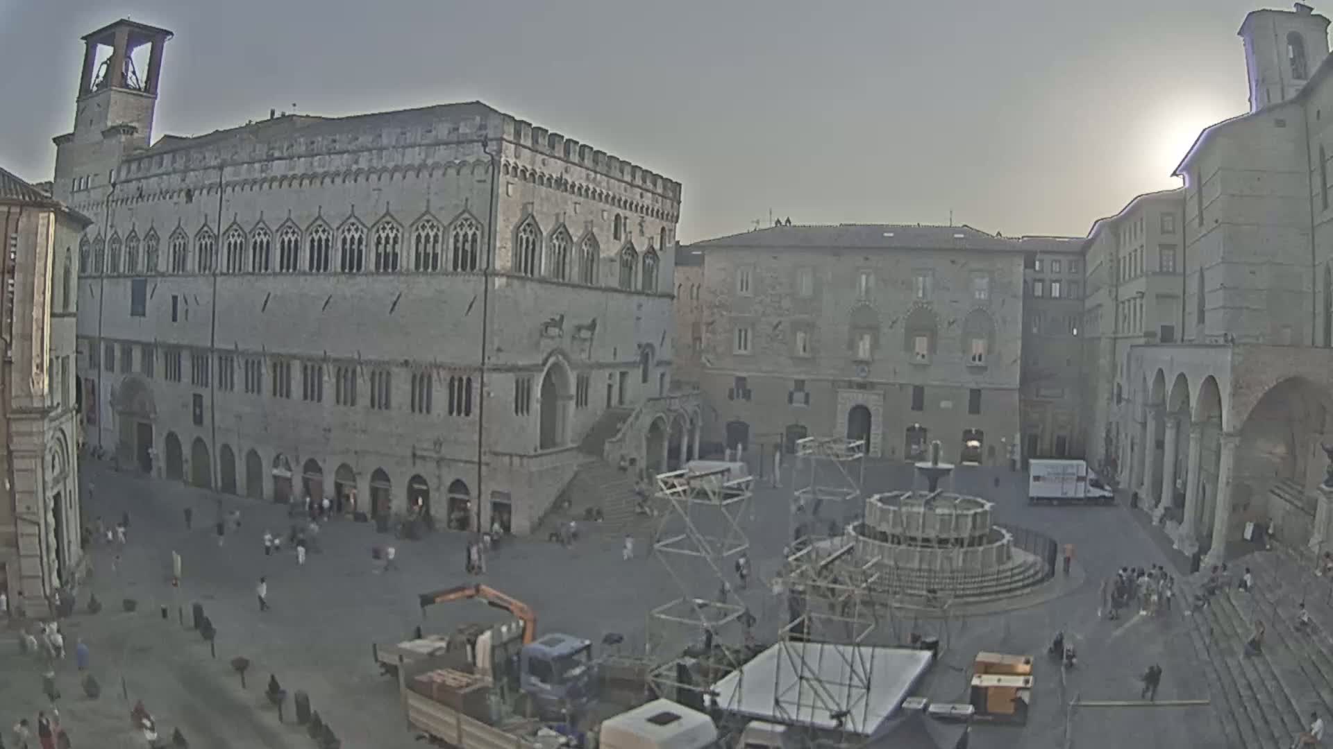 A large, stone public square with a fountain and several buildings, including a large palazzo, under a hazy, sunny sky.