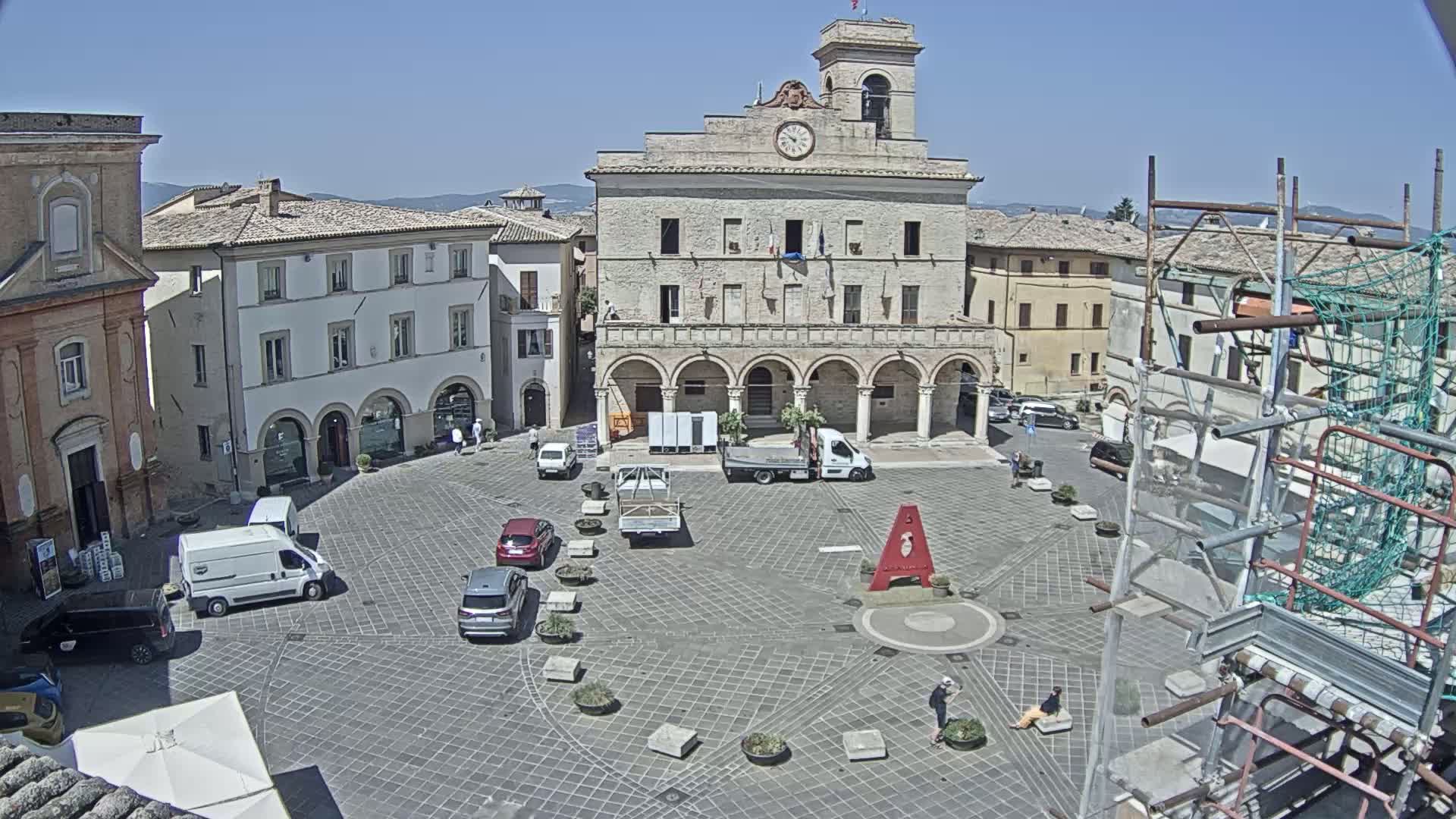 A town square with several vehicles and a large building with a clock tower in the center, under sunny skies.