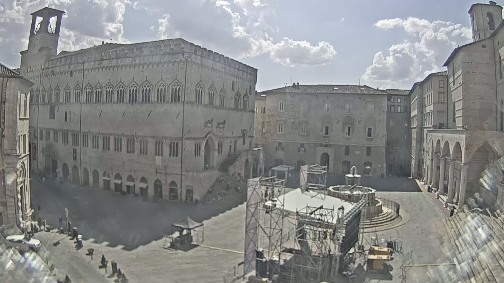 A partly sunny day reveals a large town square with historical buildings surrounding it, a fountain in the center, and scaffolding for a stage set up.