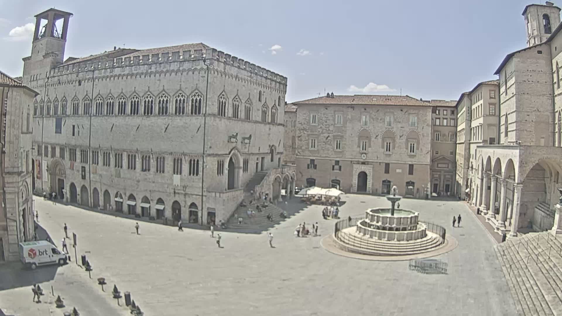 A sunny day in a large town square features several stone buildings, including a large palace with a staircase, and a central fountain.