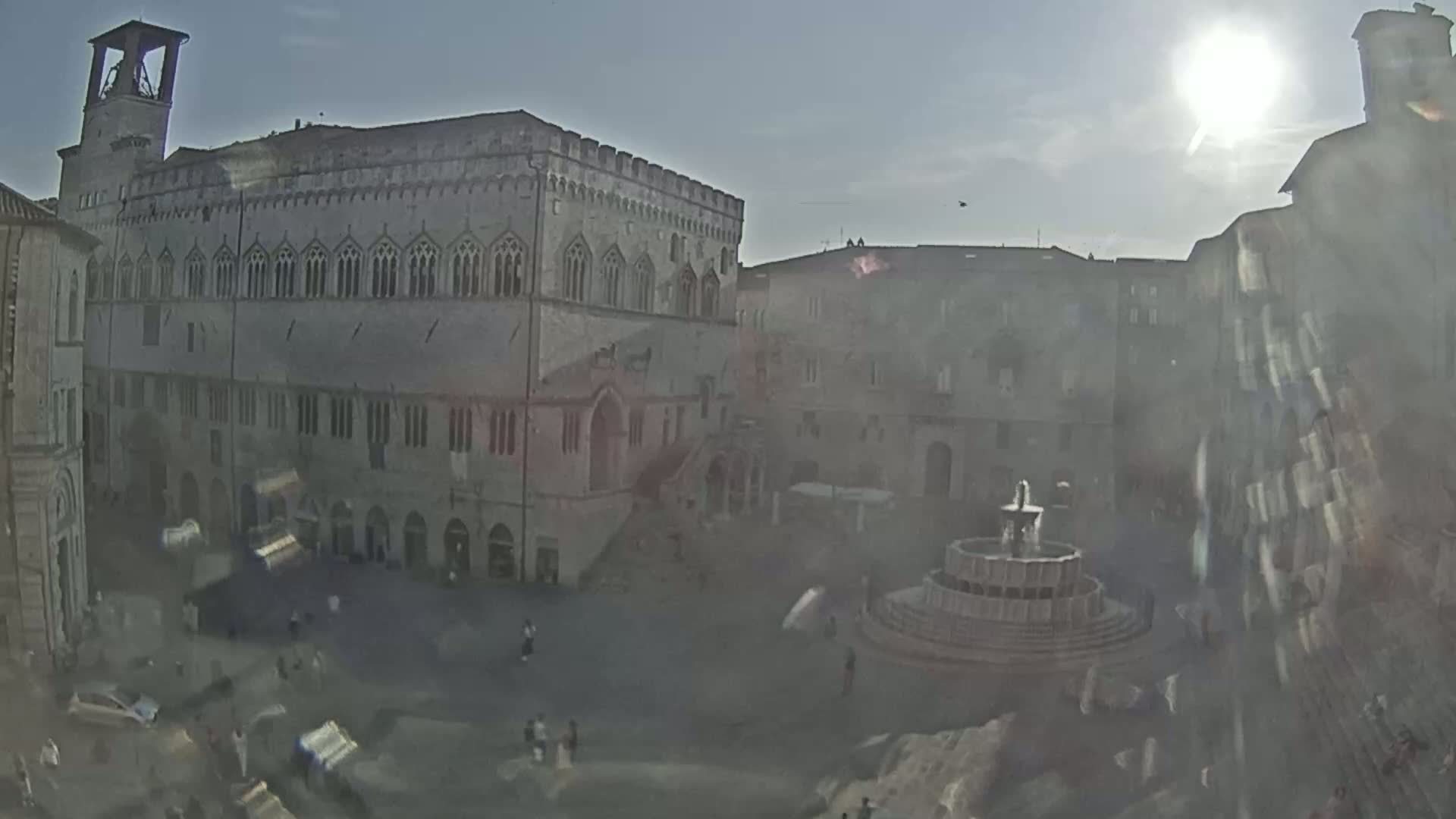 A sunny, hazy day reveals a large town square with a prominent stone building, a fountain, and several other buildings surrounding the central area where people are walking.