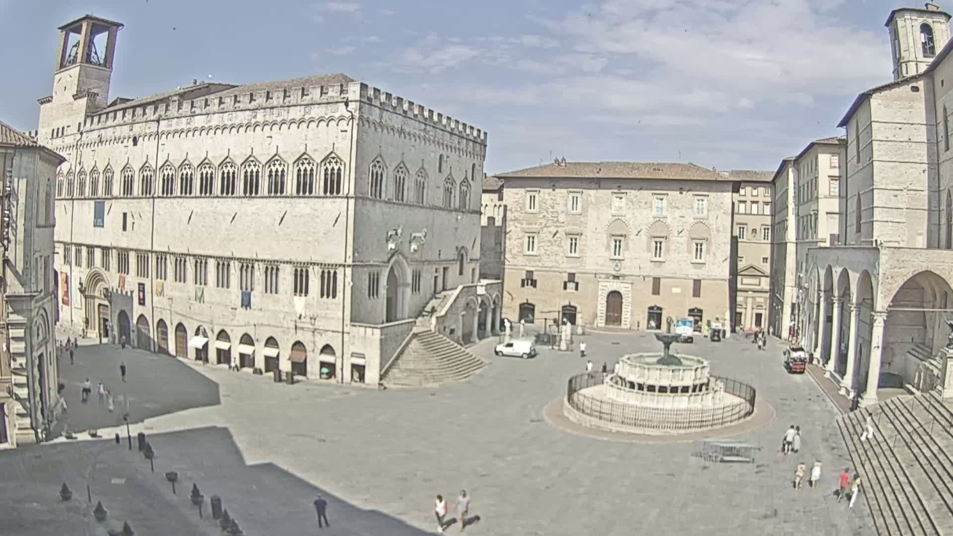 A large, mostly empty town square with a fountain in the center, surrounded by old stone buildings under a mostly sunny sky.
