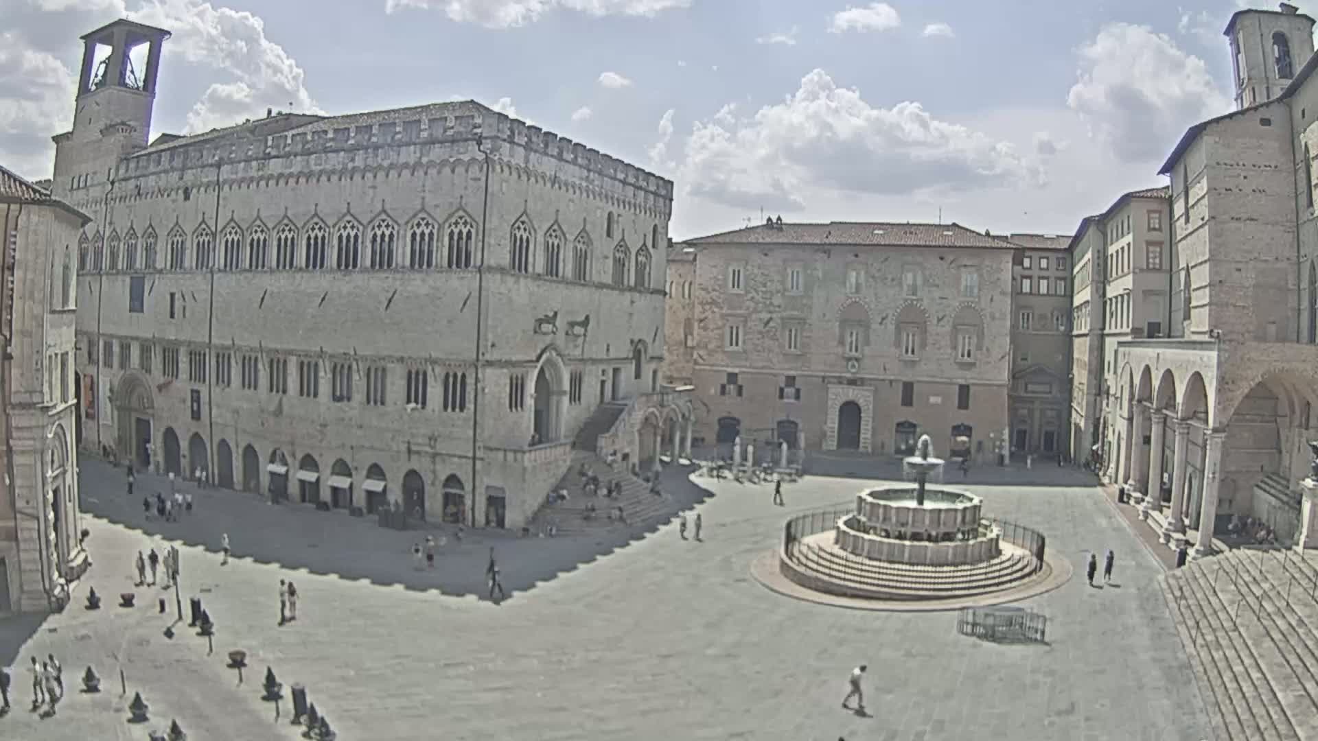 A large, stone plaza with a fountain at its center is surrounded by historic buildings under a partly cloudy sky.