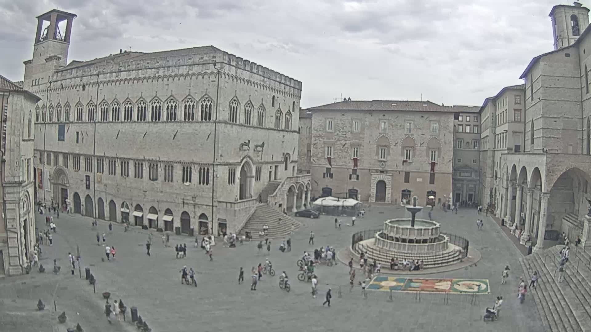 A large town square with a fountain at its center is surrounded by several stone buildings under an overcast sky.
