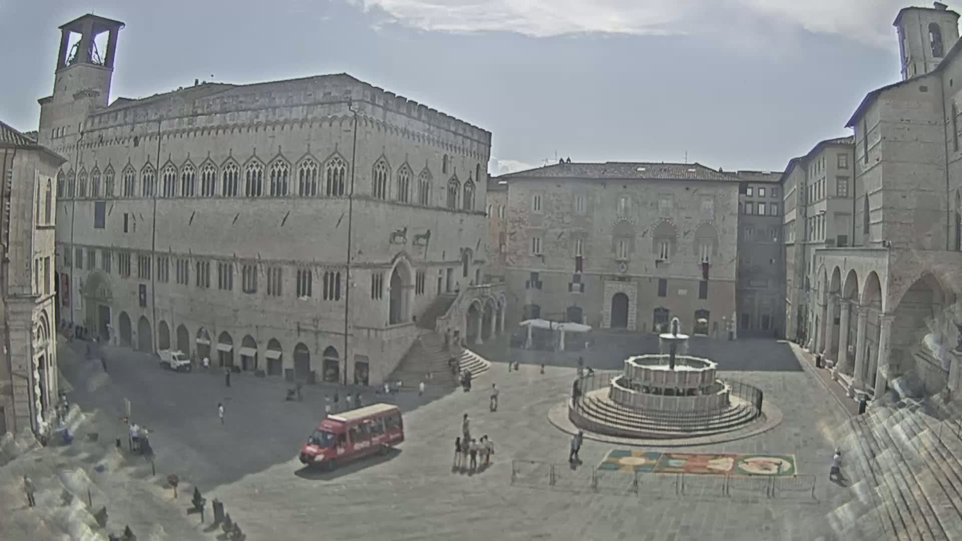 A large, stone plaza with a fountain in the center is surrounded by medieval-style buildings under a partly cloudy sky.