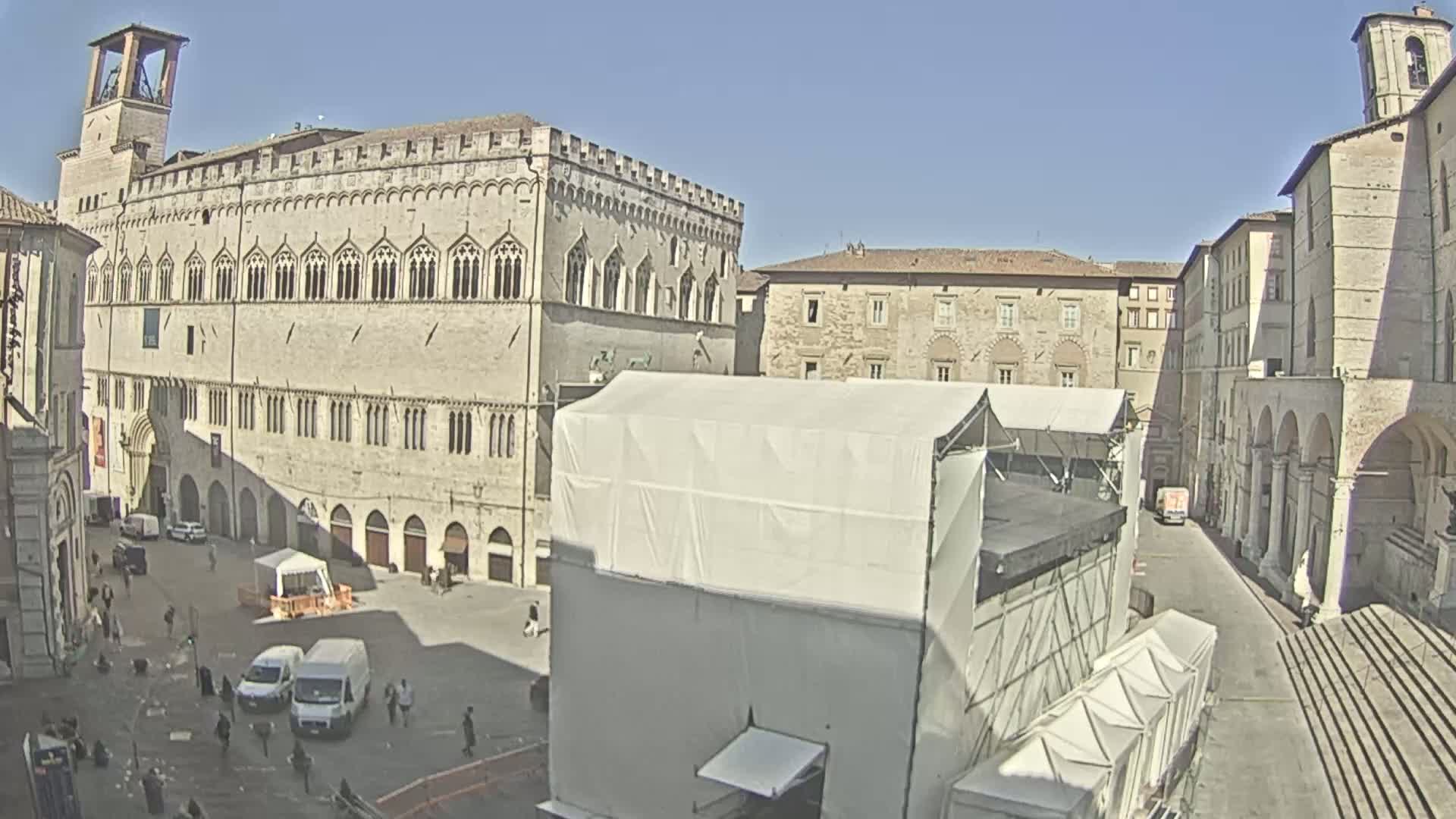 A large, stone plaza is visible, surrounded by old buildings under a clear, sunny sky; a large temporary structure dominates the center of the plaza.