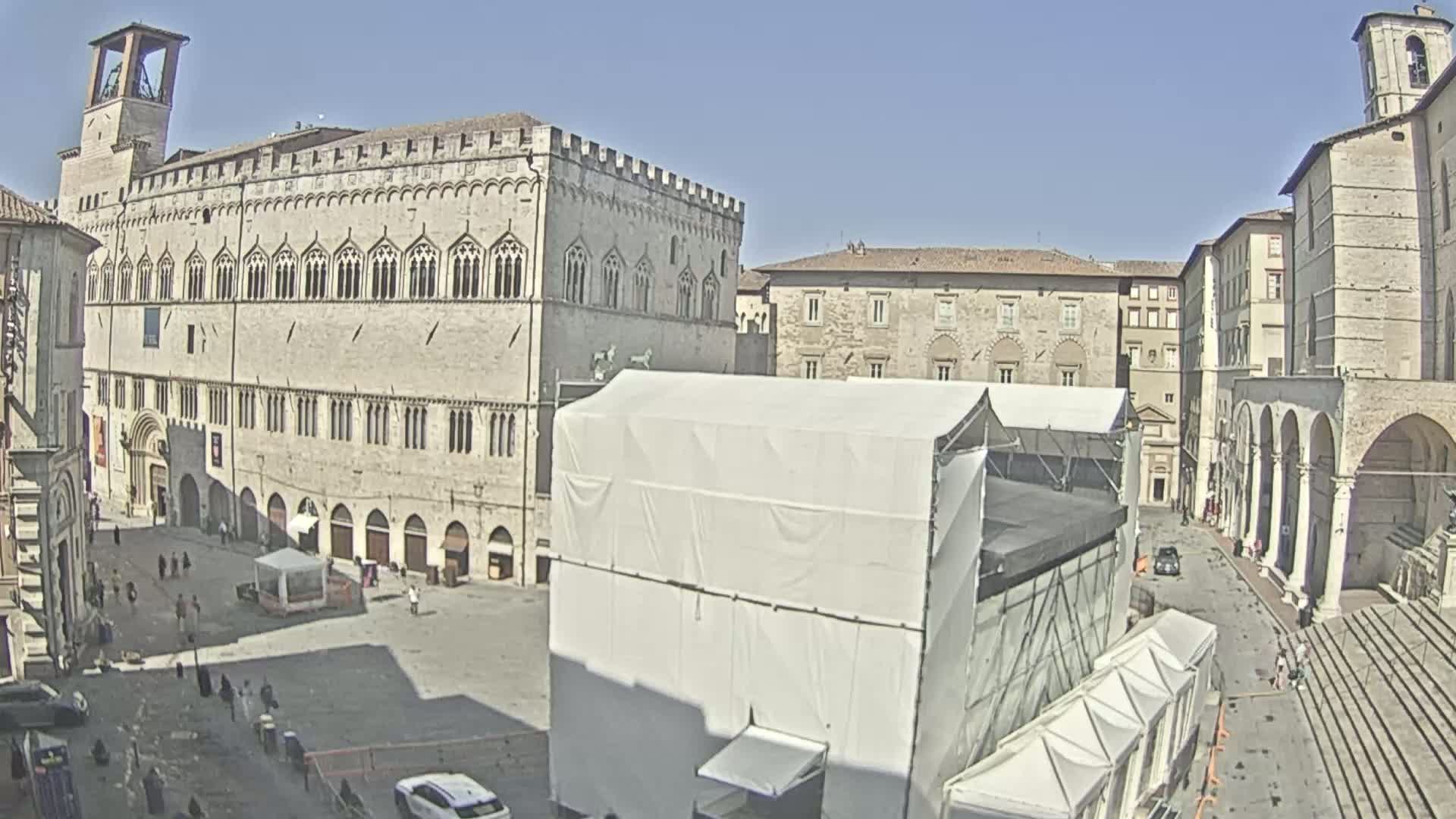 A large, light-colored stone building dominates a sunny town square, partially obscured by white temporary structures, with several smaller buildings surrounding the square and a few people scattered throughout.