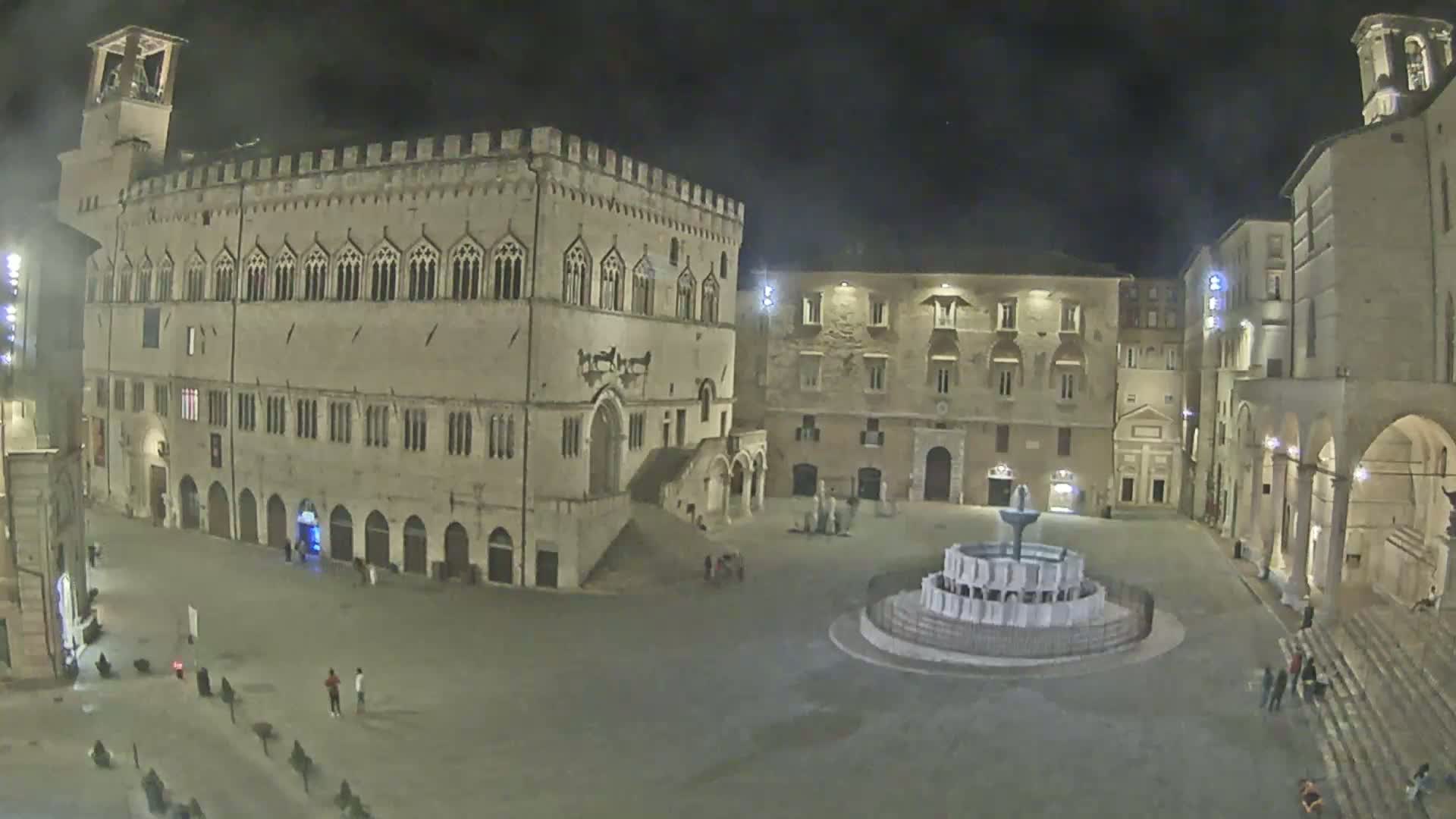 A large, light-colored stone building dominates a sunny town square, partially obscured by white temporary structures, with several smaller buildings surrounding the square and a few people scattered throughout.