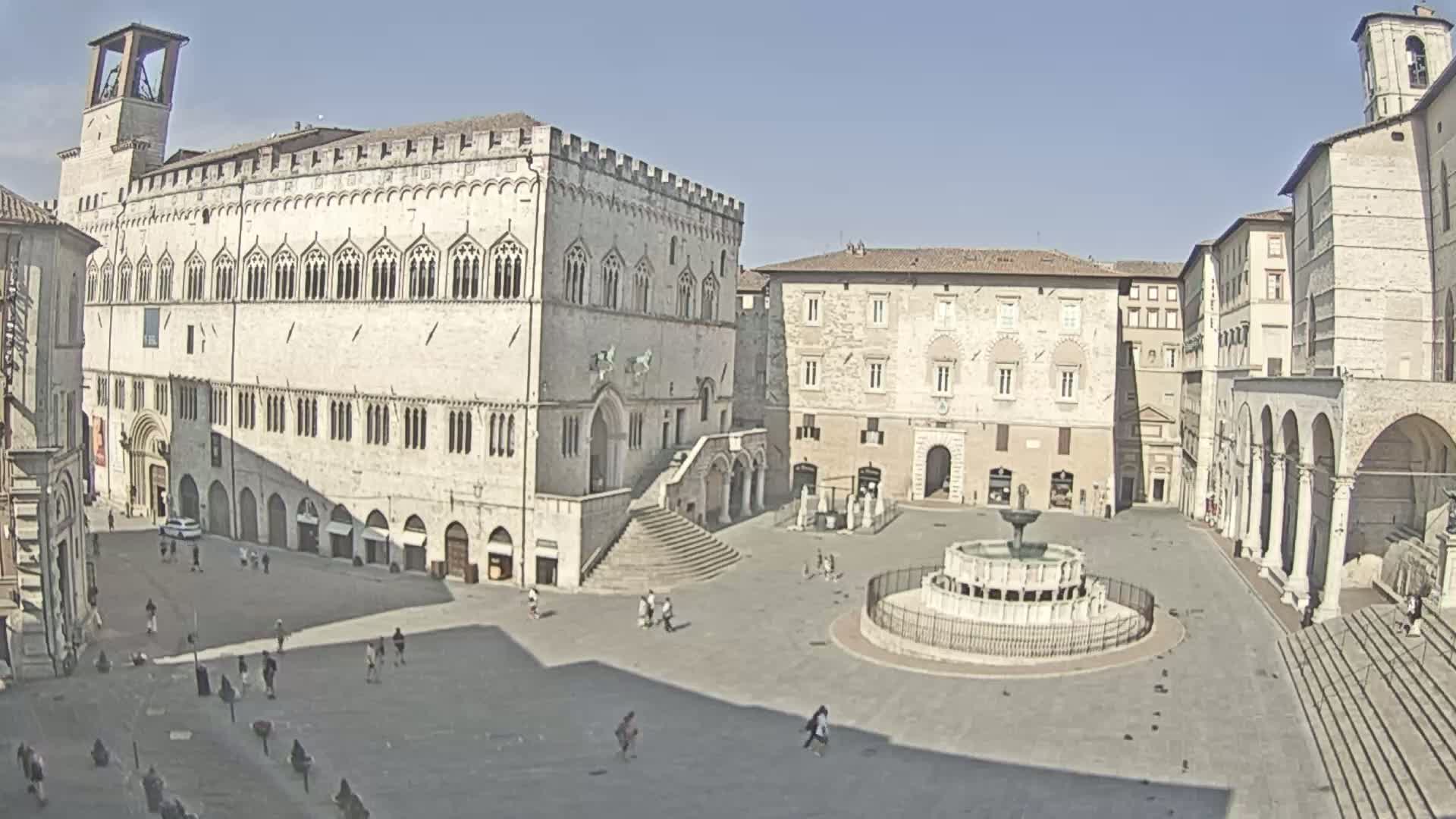 A large, mostly empty town square, under sunny skies, is surrounded by ancient stone buildings, including a large palace and a fountain in the center.