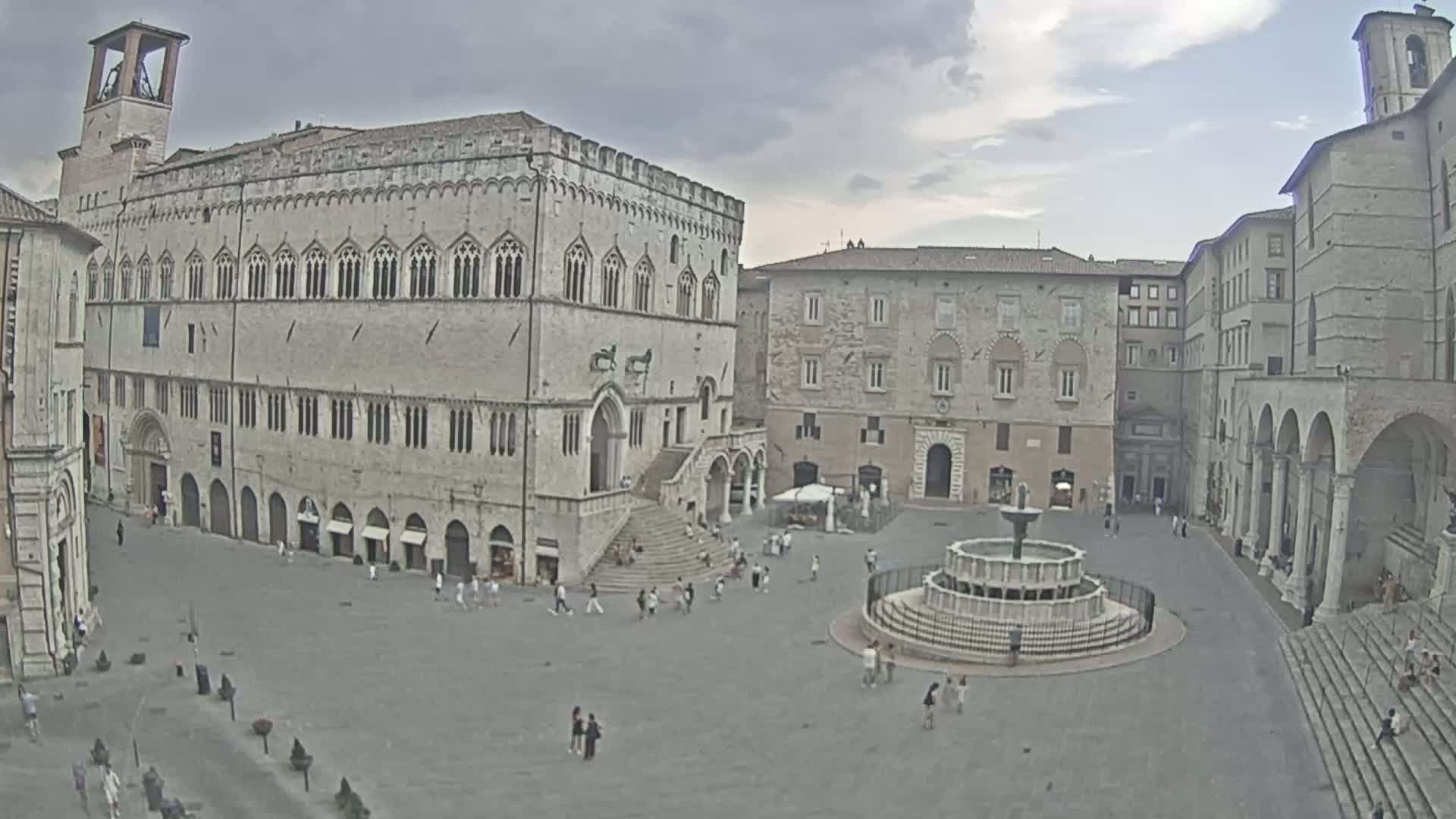 A large, mostly empty town square, under sunny skies, is surrounded by ancient stone buildings, including a large palace and a fountain in the center.