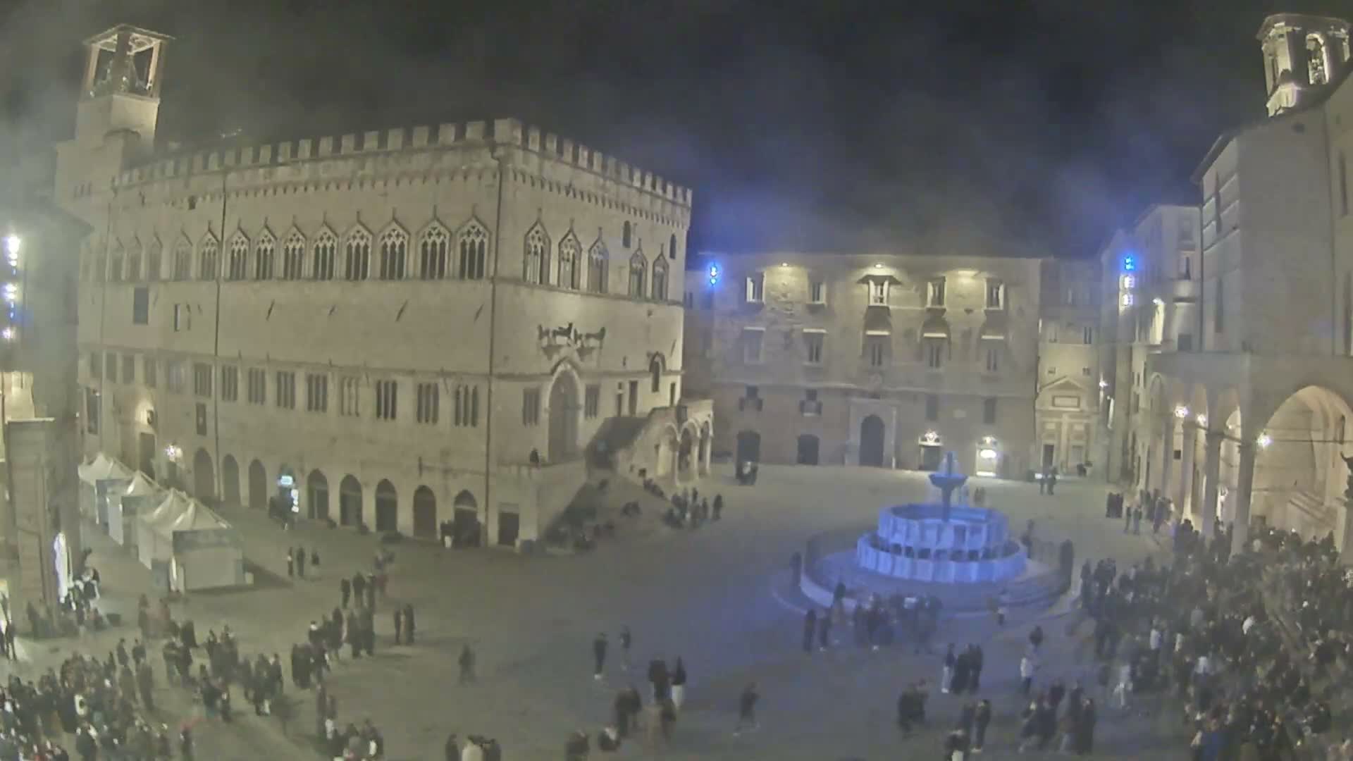 A historic town square is brightly illuminated at night, filled with numerous people gathered around a prominent blue-lit tiered fountain, flanked by grand old buildings and a row of white market tents, all under a dark, hazy sky.