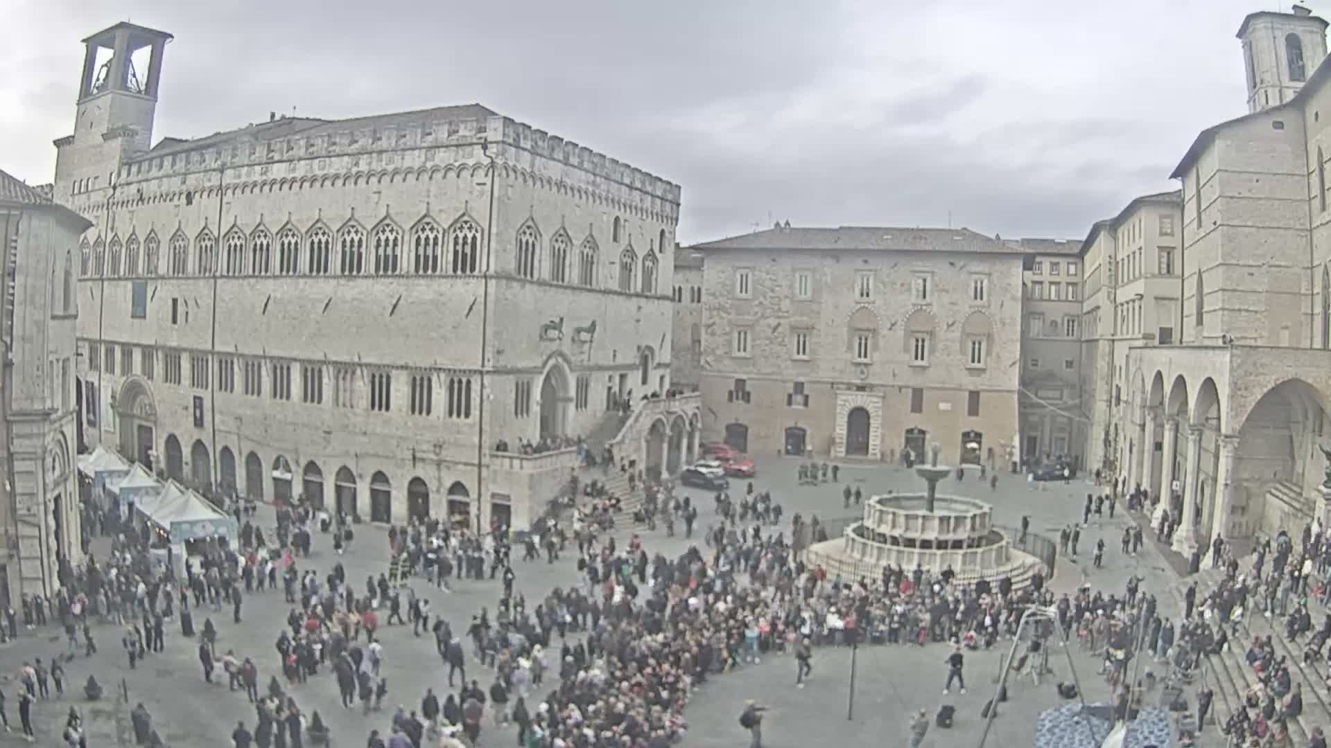 A bustling historic city square is viewed under a cloudy sky, surrounded by ornate stone buildings and a prominent bell tower, with market tents and a multi-tiered fountain amidst a large crowd of people.