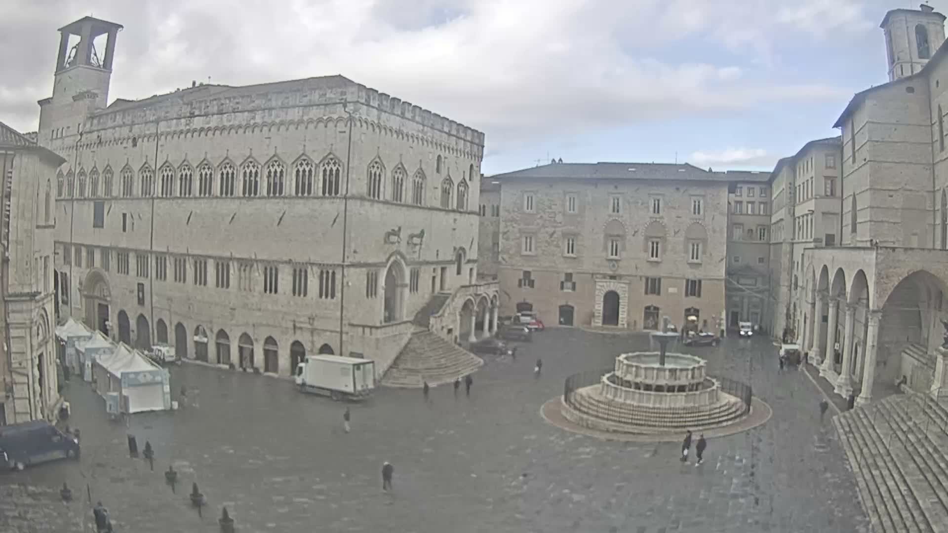 A historic European piazza, featuring a large tiered fountain and surrounded by ornate stone buildings, including a prominent bell tower and arched pathways, hosts scattered pedestrians and vehicles on a damp, overcast day.