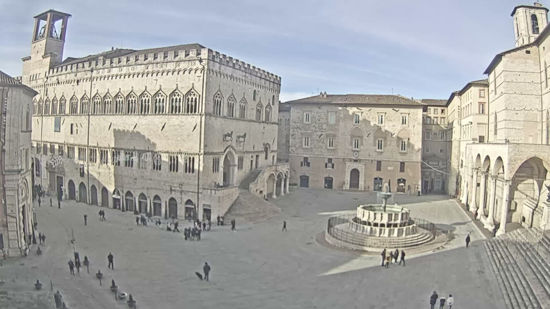 A wide-angle view captures a historic Italian piazza under a clear, sunny sky, showcasing a grand Gothic-style palace with a bell tower on the left, a prominent tiered fountain, and numerous people traversing the open square.