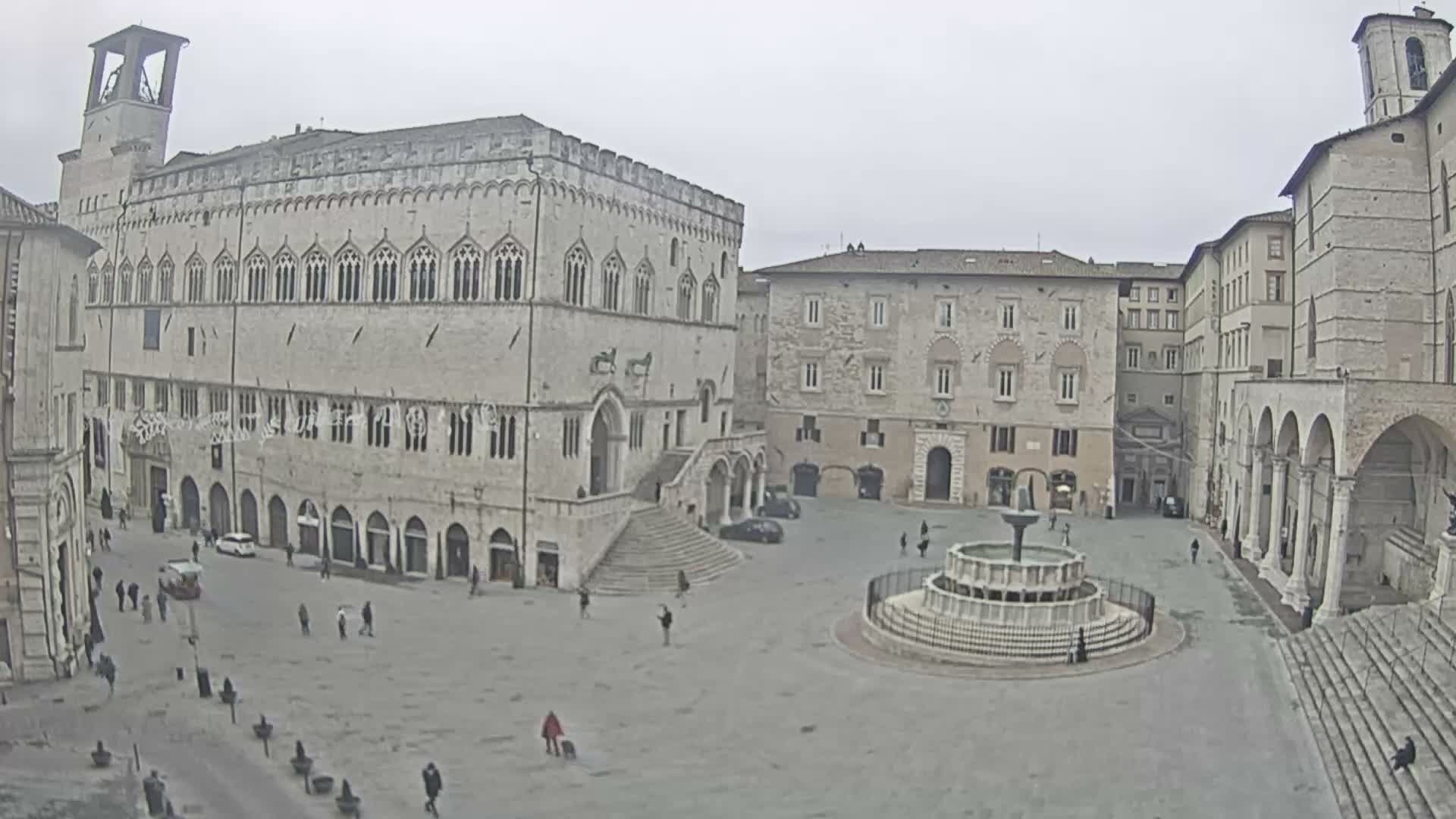 A wide view of a grand medieval Italian piazza features a prominent Gothic palace with a bell tower, a large ornate fountain, and scattered pedestrians under a uniformly overcast sky.