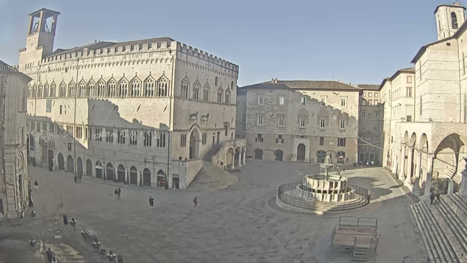 A wide view of a grand medieval Italian piazza features a prominent Gothic palace with a bell tower, a large ornate fountain, and scattered pedestrians under a uniformly overcast sky.