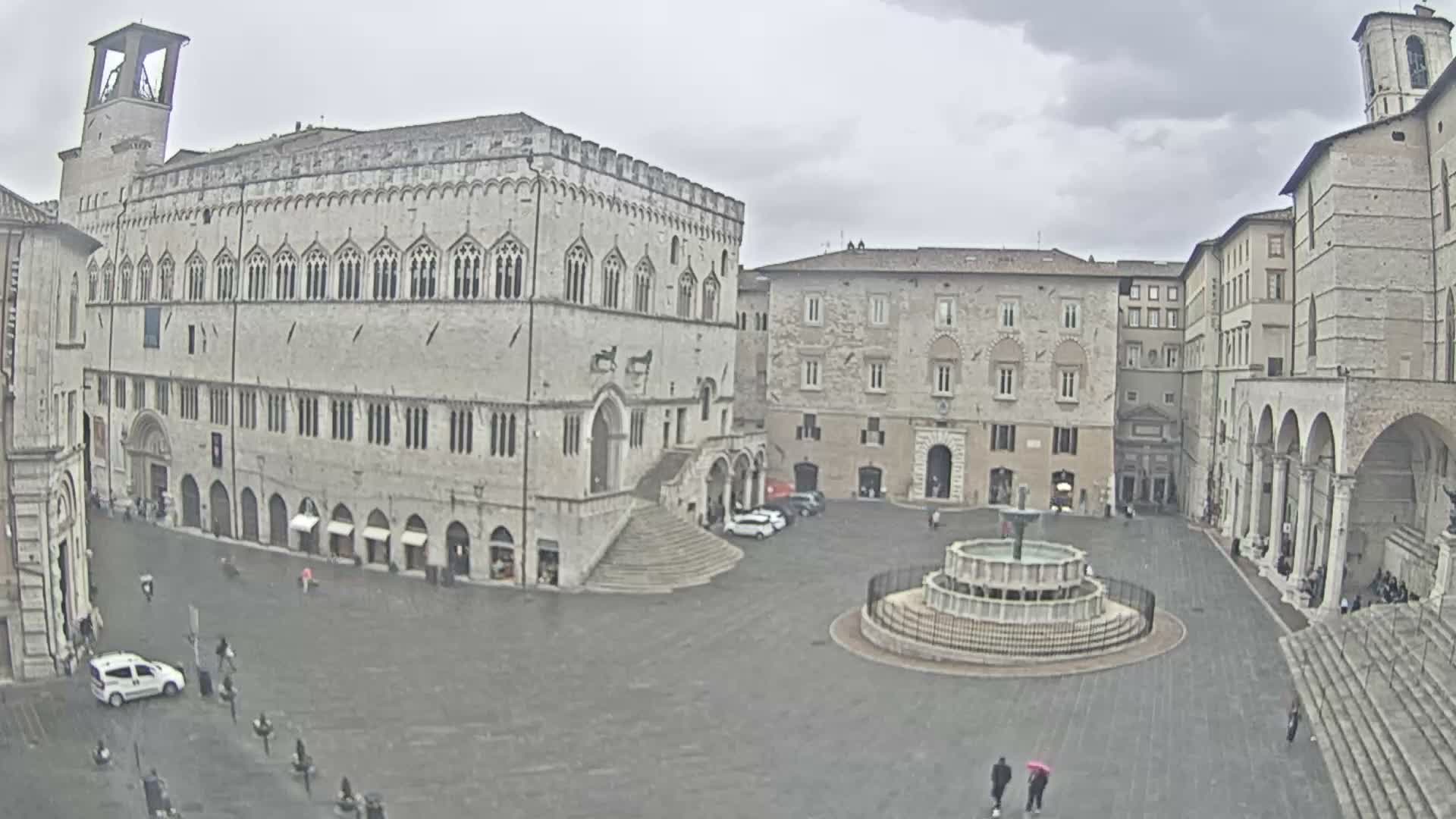 A wide view of a grand medieval Italian piazza features a prominent Gothic palace with a bell tower, a large ornate fountain, and scattered pedestrians under a uniformly overcast sky.