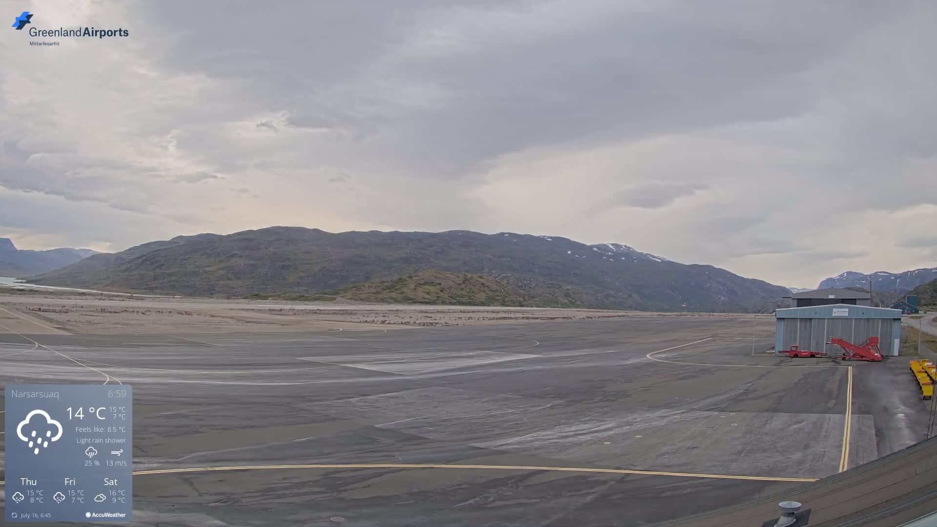 A mostly empty airport tarmac is shown under a light rain shower, with mountains in the background.