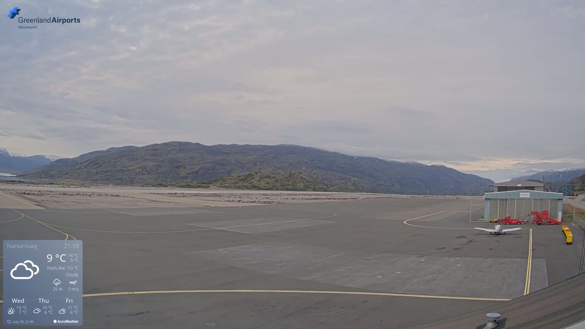 A small airplane sits on a tarmac at an airport surrounded by low mountains under a partly cloudy sky.