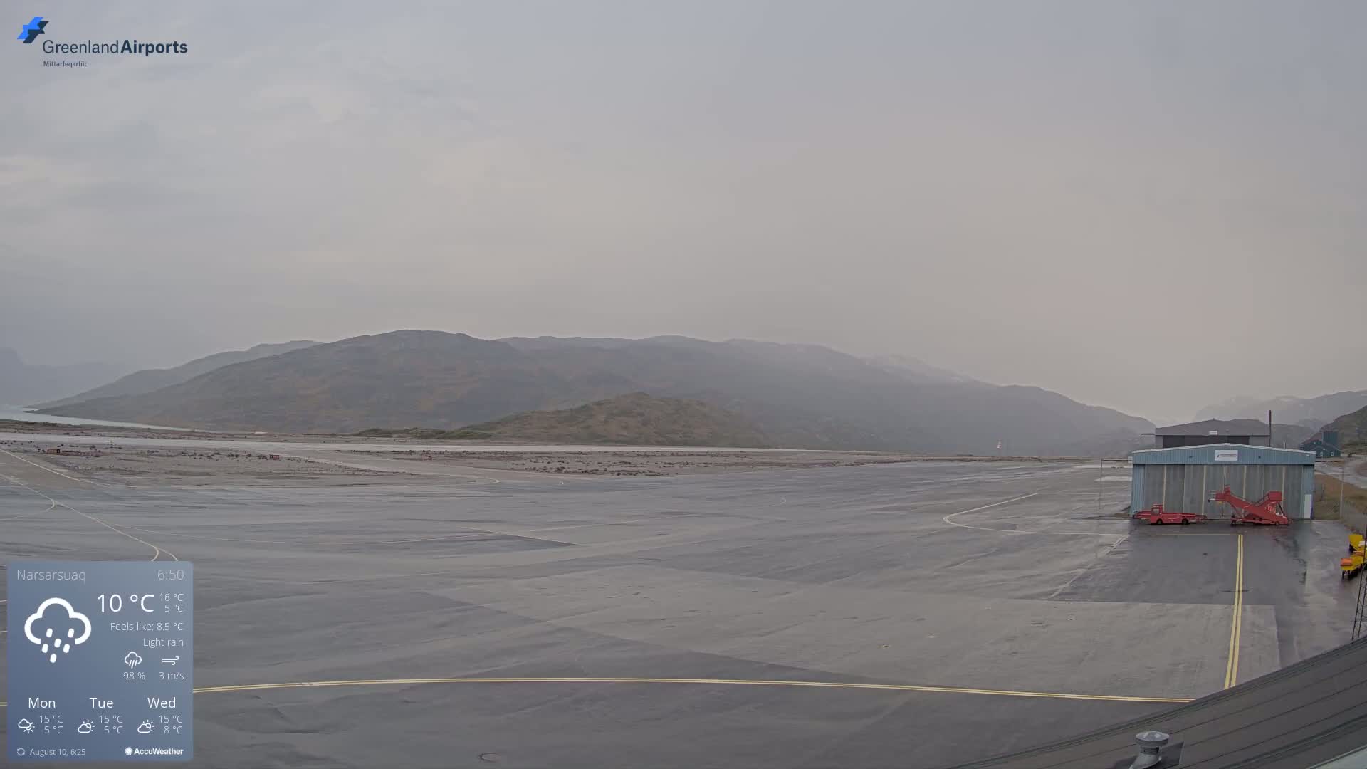 A wet airport runway with a hangar and ground support equipment is visible under a light rain and hazy sky, with mountains in the background.