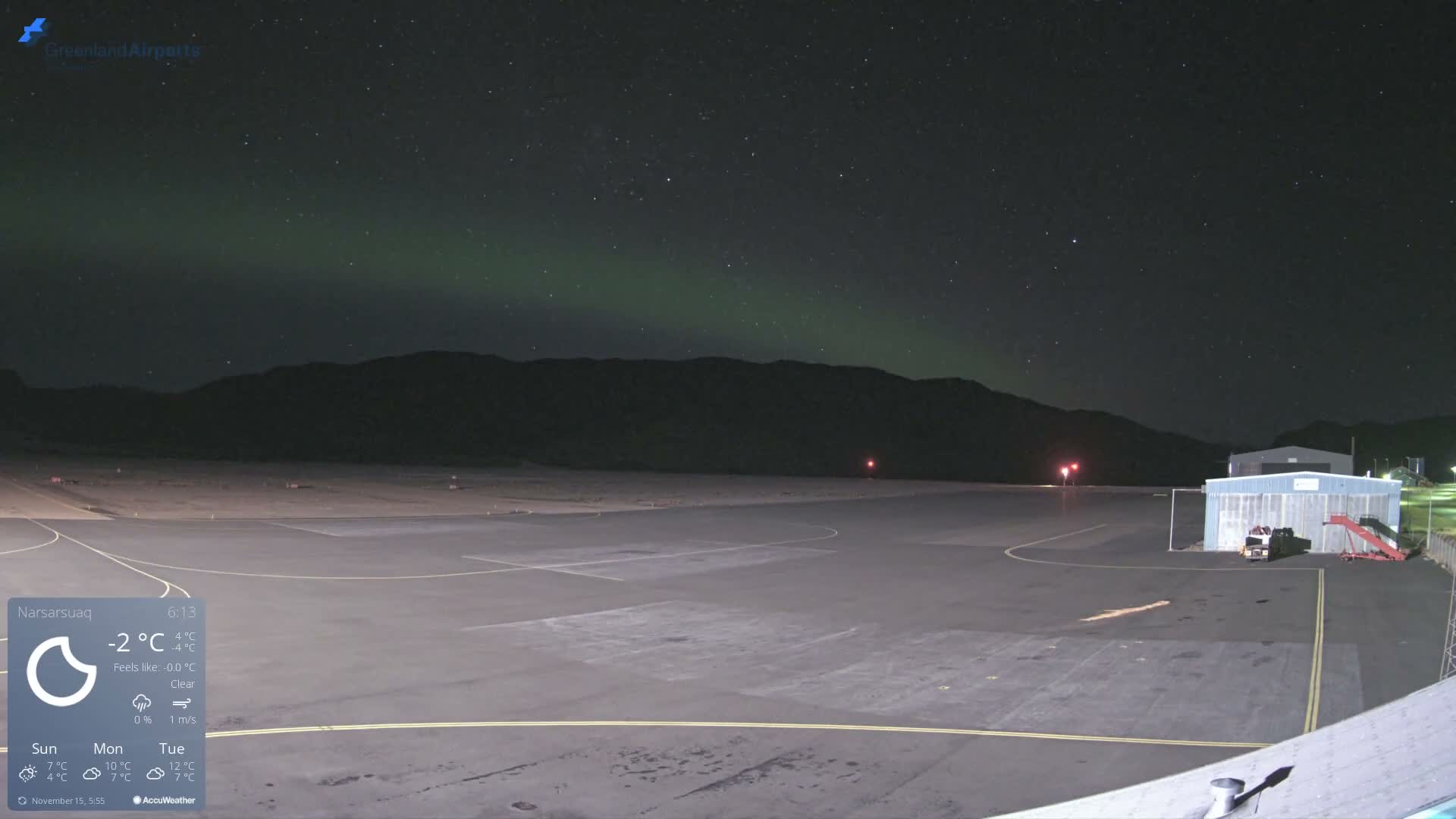 An illuminated airport tarmac and buildings are seen under a clear, star-filled night sky with a green aurora borealis, framed by distant mountains, with the temperature at -2°C and clear conditions.