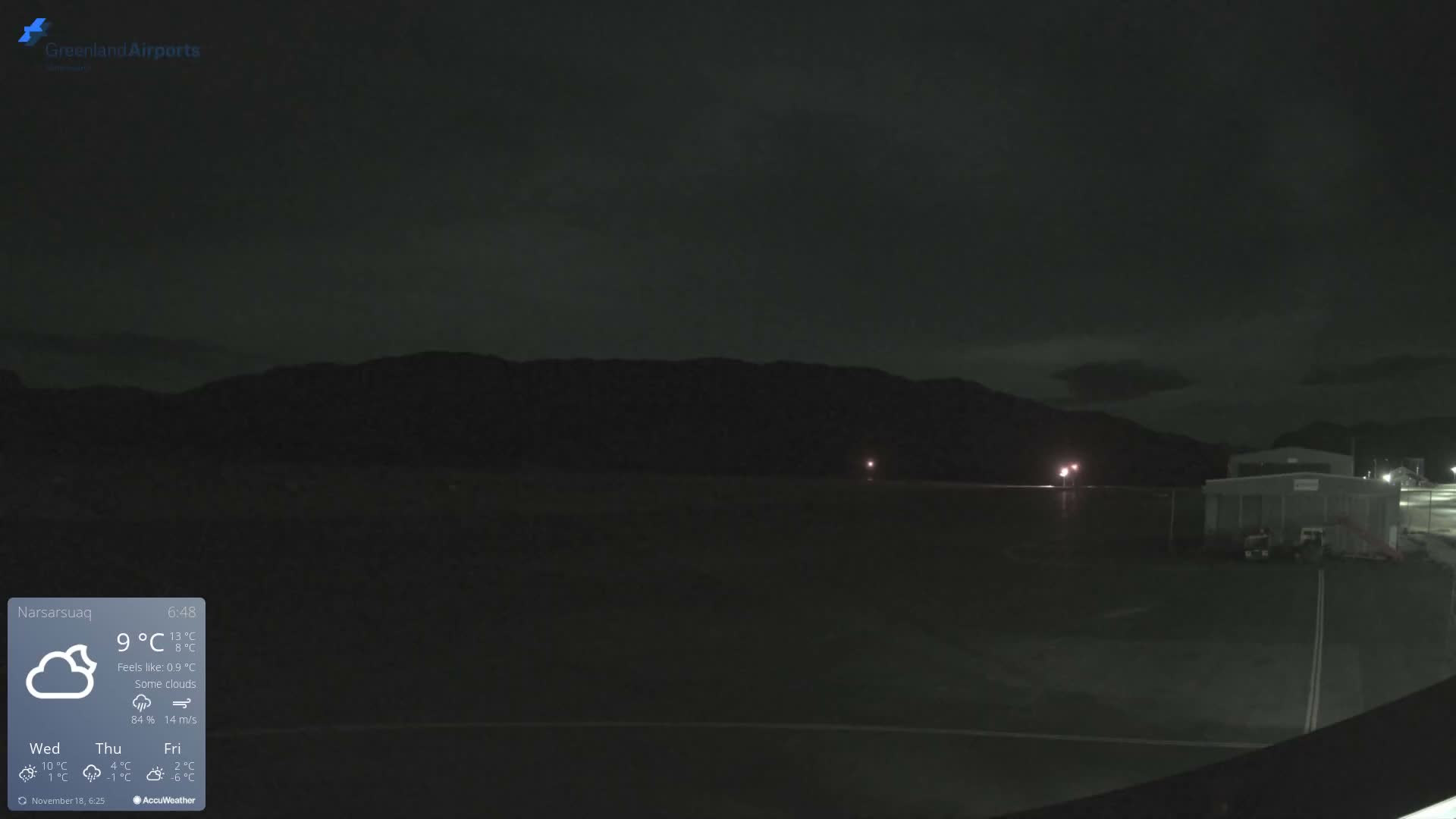 The image captures a dark, cloudy night view of an airfield, with distant mountains silhouetted against the horizon and dimly lit buildings beside a flat runway surface in the foreground.