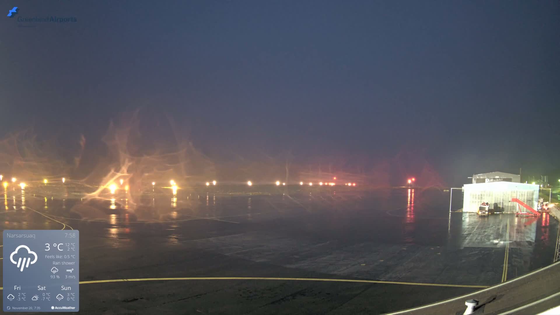 A dark airport tarmac is illuminated by numerous bright lights reflecting on its wet surface under rainy conditions.