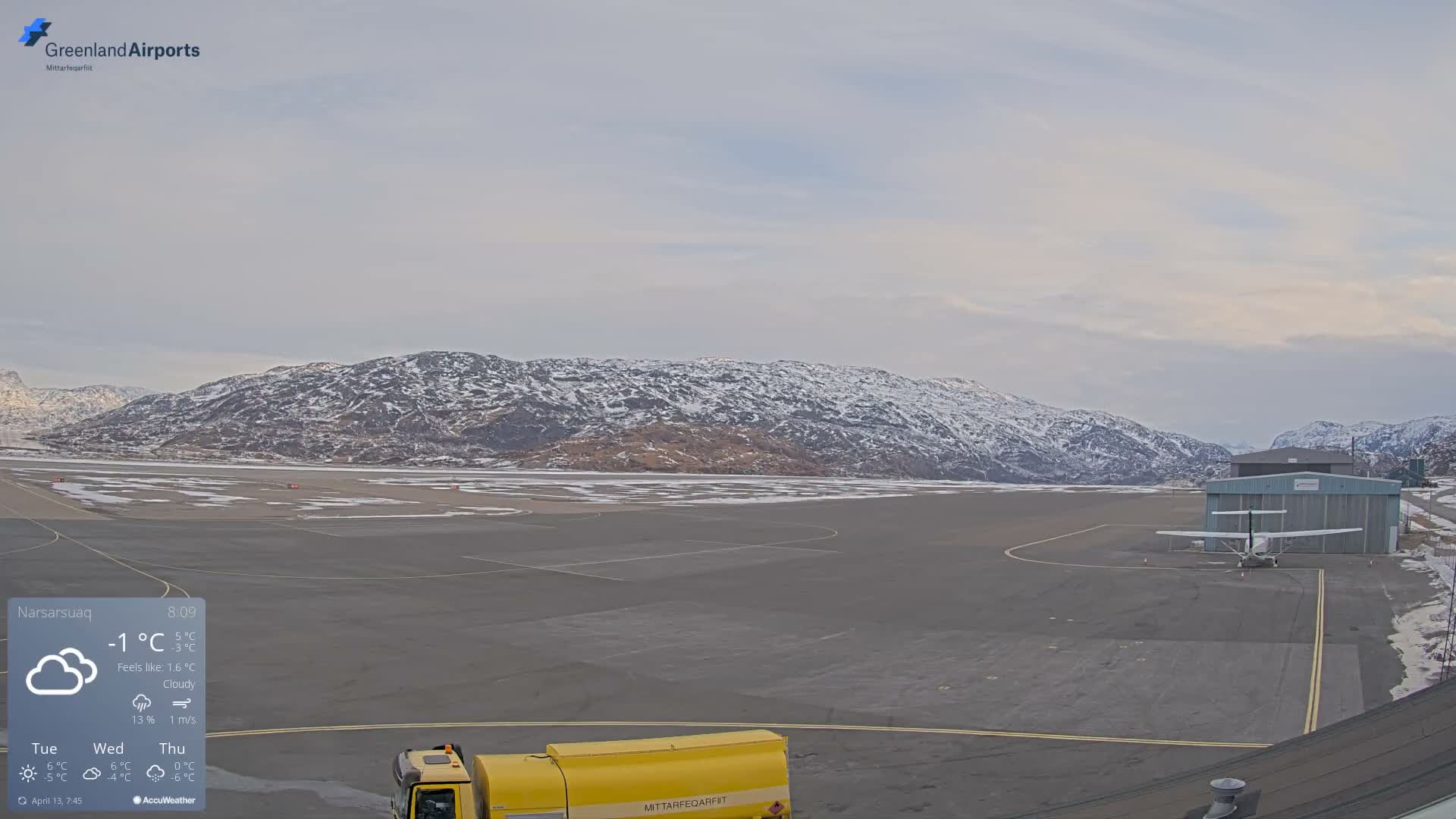 The cold, partially cloudy night sky, dotted with stars, hangs over a vast snow-covered airfield flanked by dark mountains, with distant red lights glowing and low buildings visible on the right.