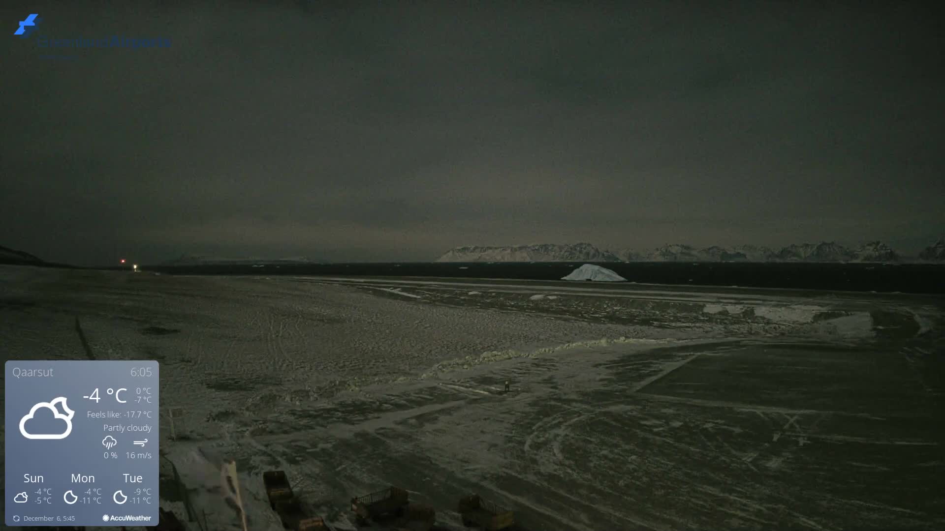 The image displays a vast, dark, snow and ice-covered landscape with distant airfield lights, a solitary person, and a body of water featuring an iceberg, all set against a backdrop of snow-capped mountains under a cold, overcast sky.