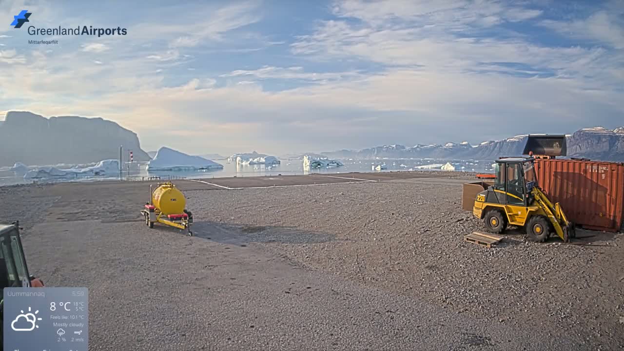 A gravel airfield with a small yellow loader and a fuel tanker sits near a body of water dotted with icebergs under a mostly cloudy sky.