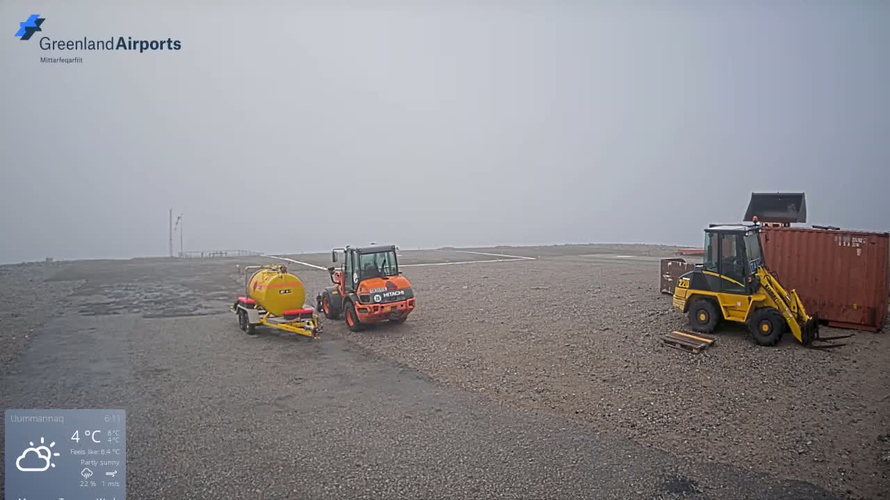 A foggy airfield with two small construction vehicles, a fuel tanker, and a shipping container parked on a gravel surface.