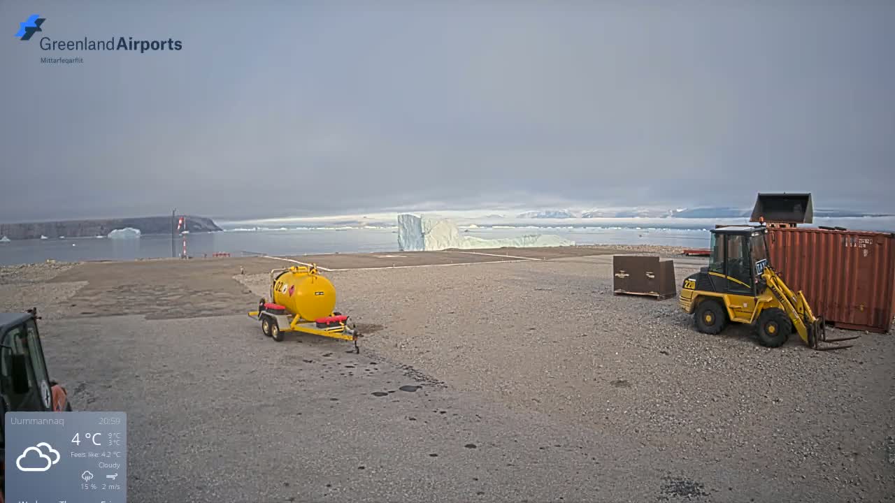 A foggy airfield with two small construction vehicles, a fuel tanker, and a shipping container parked on a gravel surface.