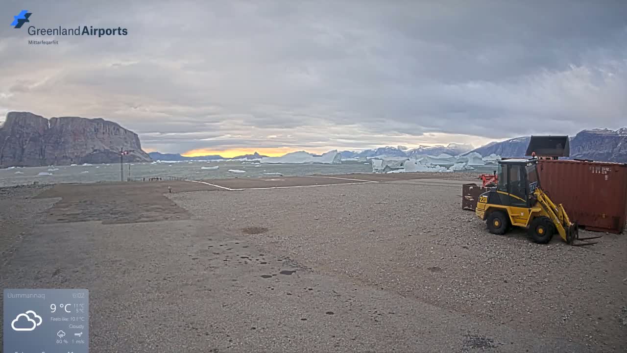 A small yellow loader sits near a shipping container in a gravel lot overlooking a calm body of water with icebergs and a rocky shore under a cloudy sky.