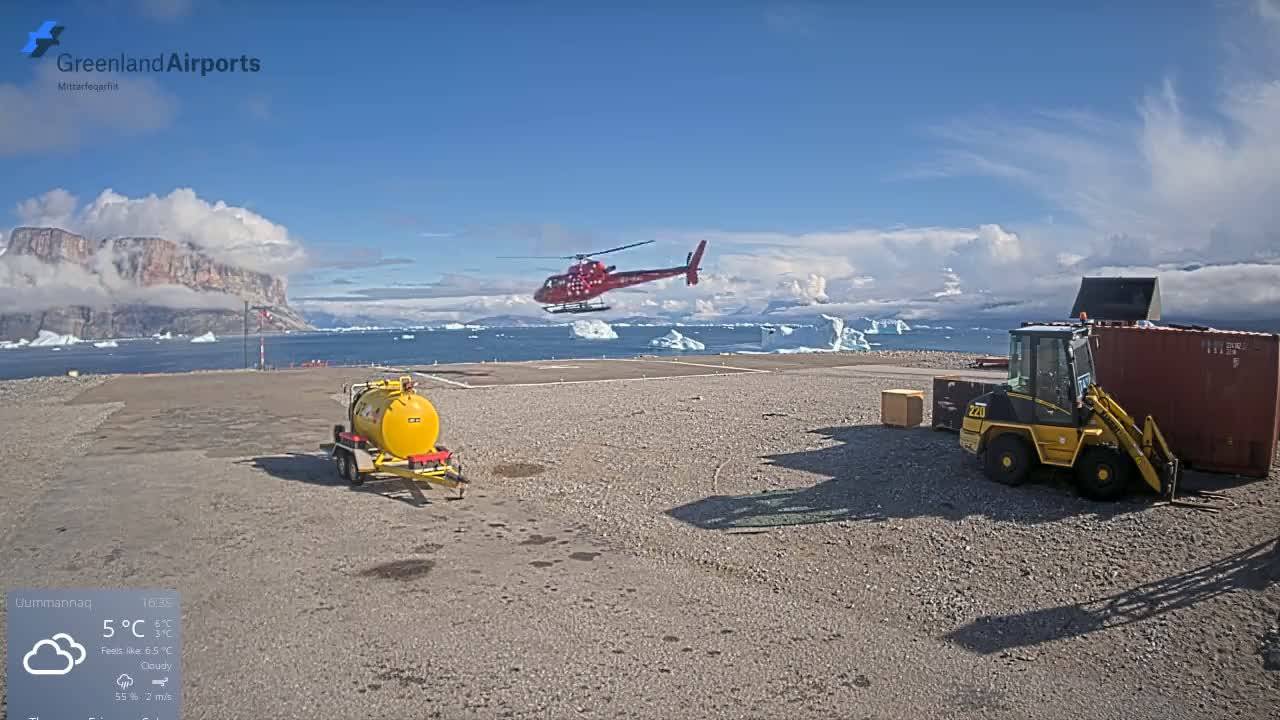 A red helicopter is hovering over a helipad near a body of water with icebergs, under a partly cloudy sky.