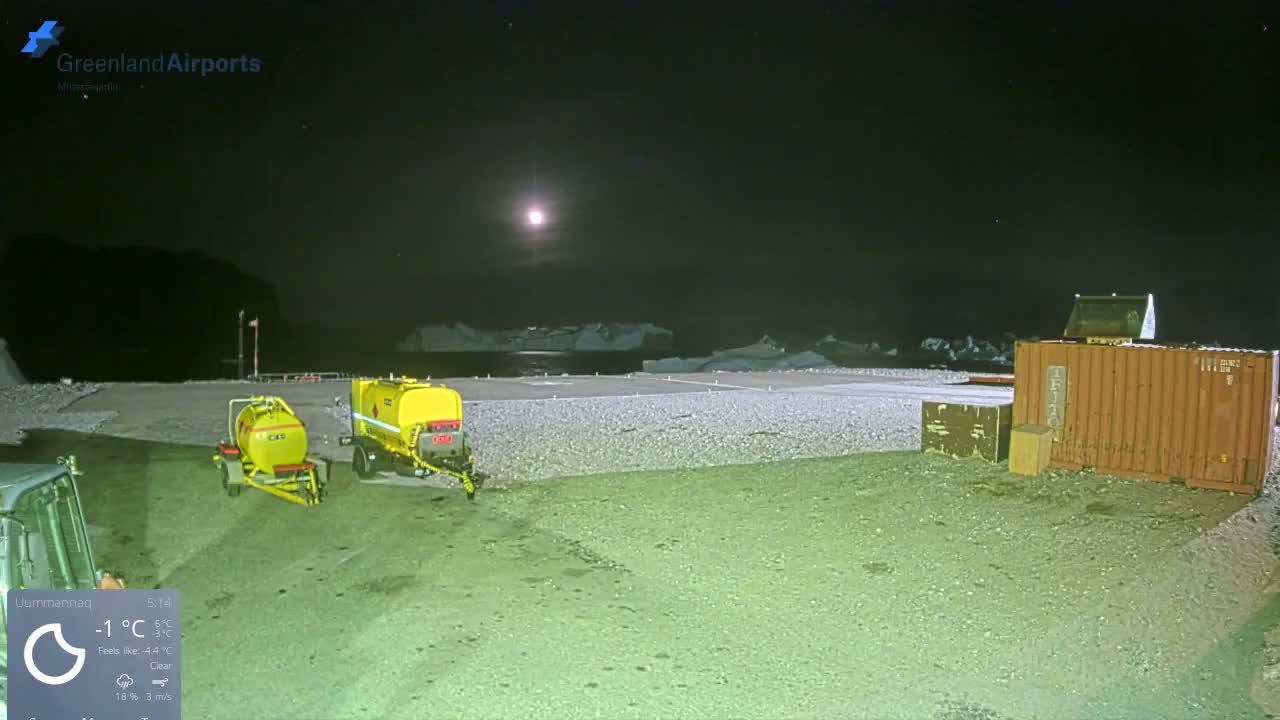 An outdoor industrial area or airport at night features two yellow tank trailers and a large shipping container on a gravelly ground, with a full moon illuminating icebergs in the distant water under clear, cold conditions at -1°C.