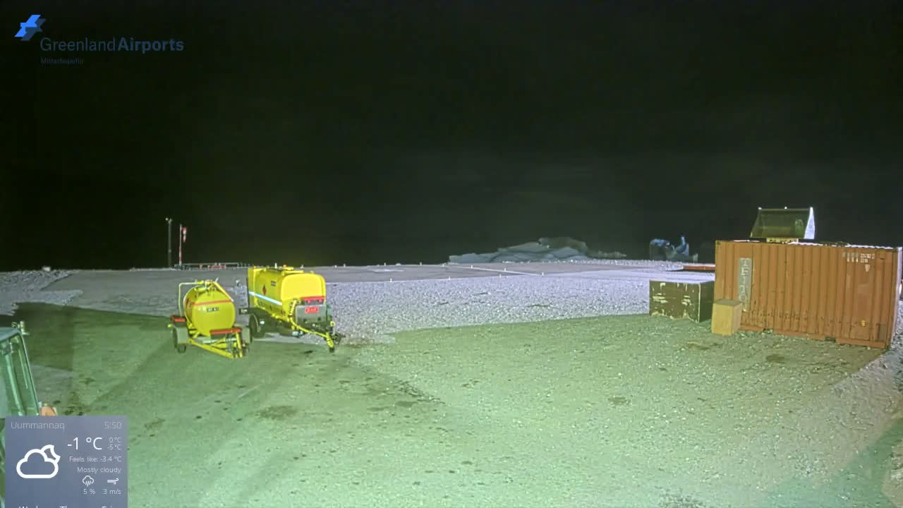Two yellow tank trailers and a large reddish-brown shipping container sit on a dark gravel and snow-dusted ground at night, with an airfield runway and distant ice-covered landforms visible under a dark, presumably cloudy sky.
