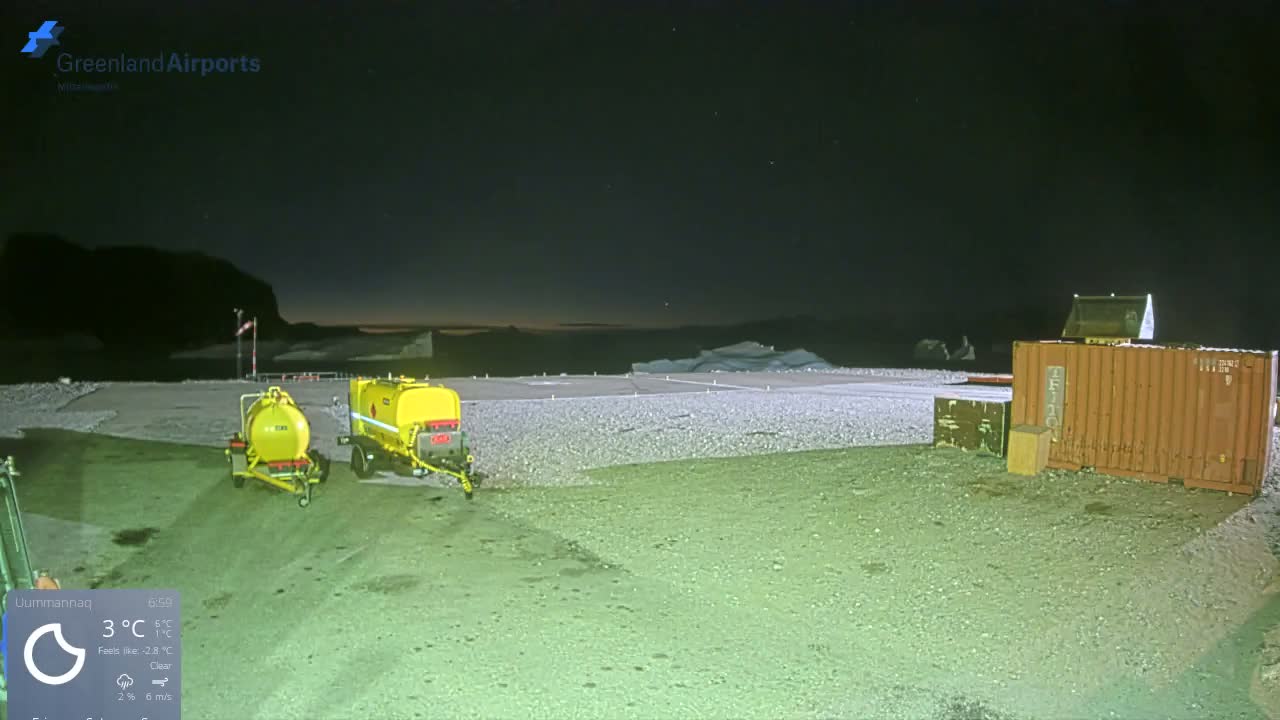 Two yellow fuel tanks and an orange shipping container sit on a gravel area next to an arctic airport runway at night, with distant icebergs and dark mountains visible under a clear, cold sky.
