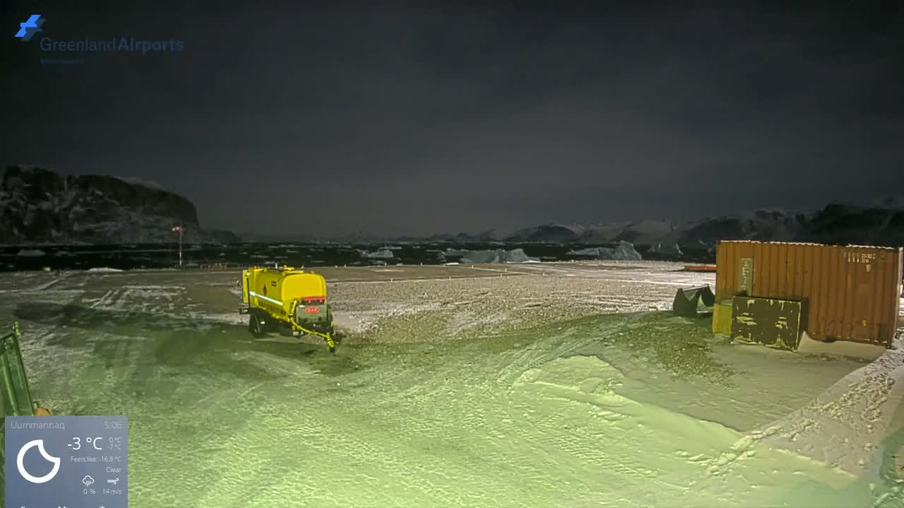 A snow-dusted airport tarmac at night, illuminated by artificial light and featuring a yellow tanker and an orange shipping container, is set against a backdrop of dark mountains and icebergs on a clear, cold evening.