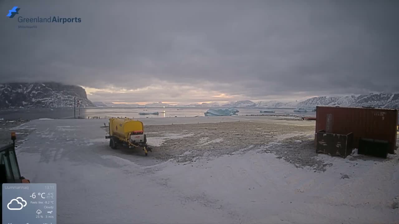 A snow-dusted airport tarmac at night, illuminated by artificial light and featuring a yellow tanker and an orange shipping container, is set against a backdrop of dark mountains and icebergs on a clear, cold evening.